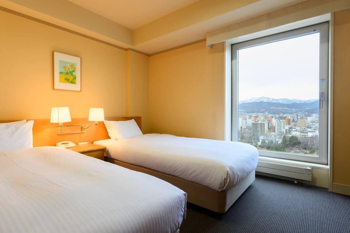 Habitación de hotel con dos camas, una mesita de noche con lámparas, un teléfono y ventana con vista a la ciudad.