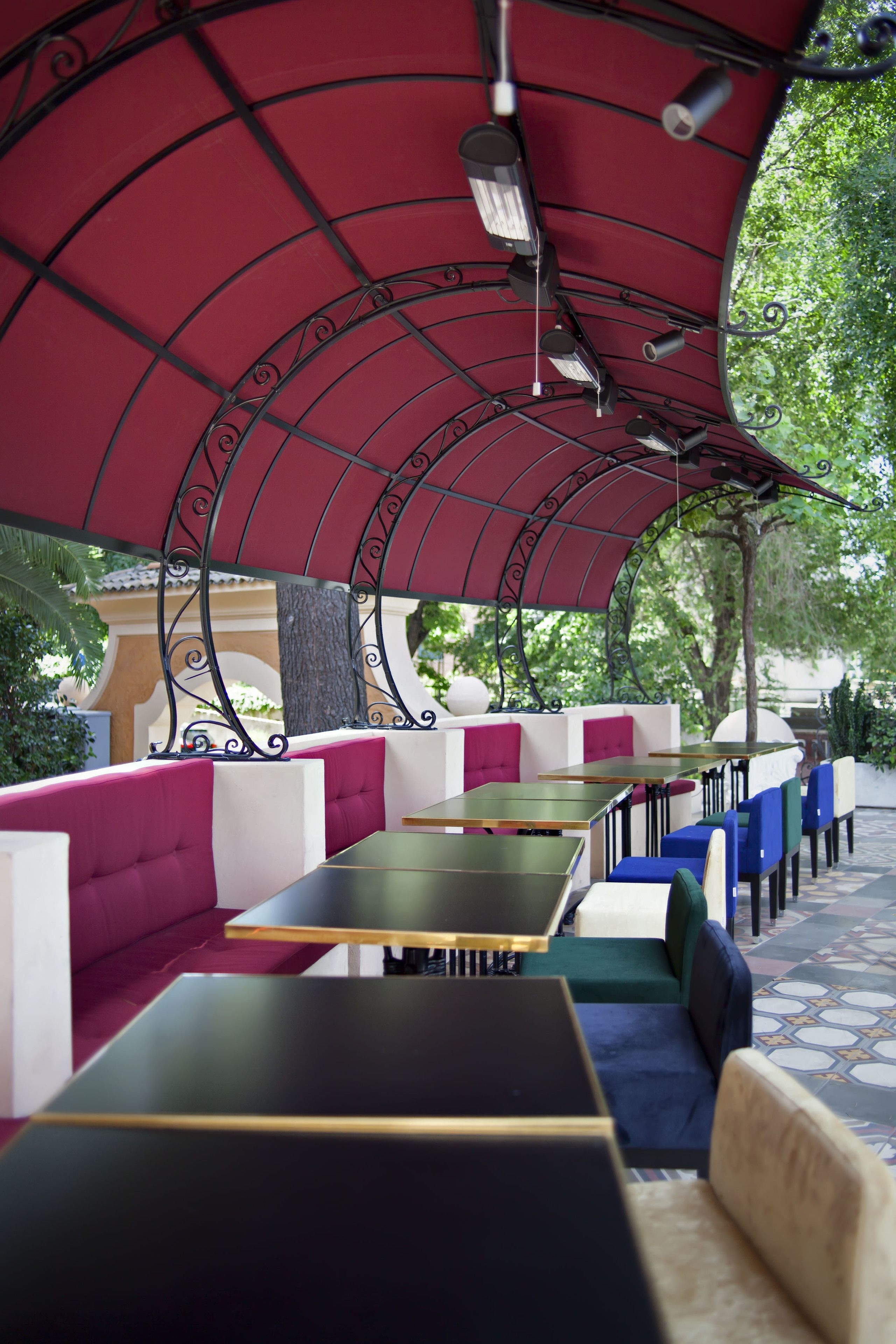 Un área de estar al aire libre con mesas y bancos acolchados, cubierta por un toldo rojo con barandillas de metal.