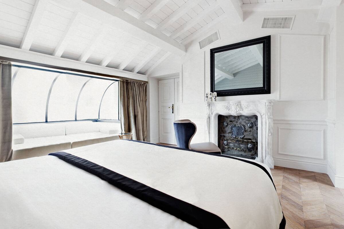 Une chambre spacieuse avec un grand lit, une cheminée et une vue sur l’extérieur, visible à travers une fenêtre ornée de rideaux.