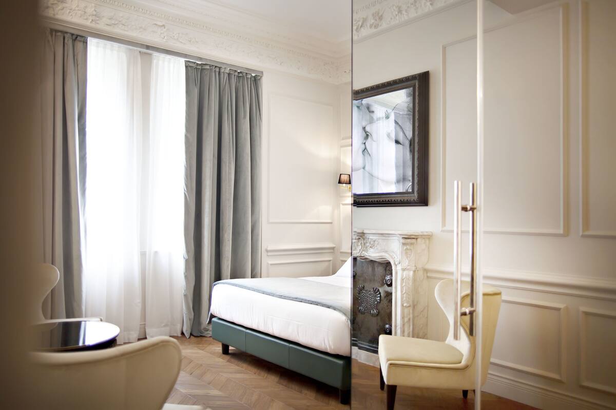 Une chambre d’hôtel avec un lit, une chaise, une cheminée et une grande fenêtre avec des rideaux.