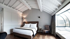 A bedroom with a large bed, a chair, a small table, and a window with a view of buildings.