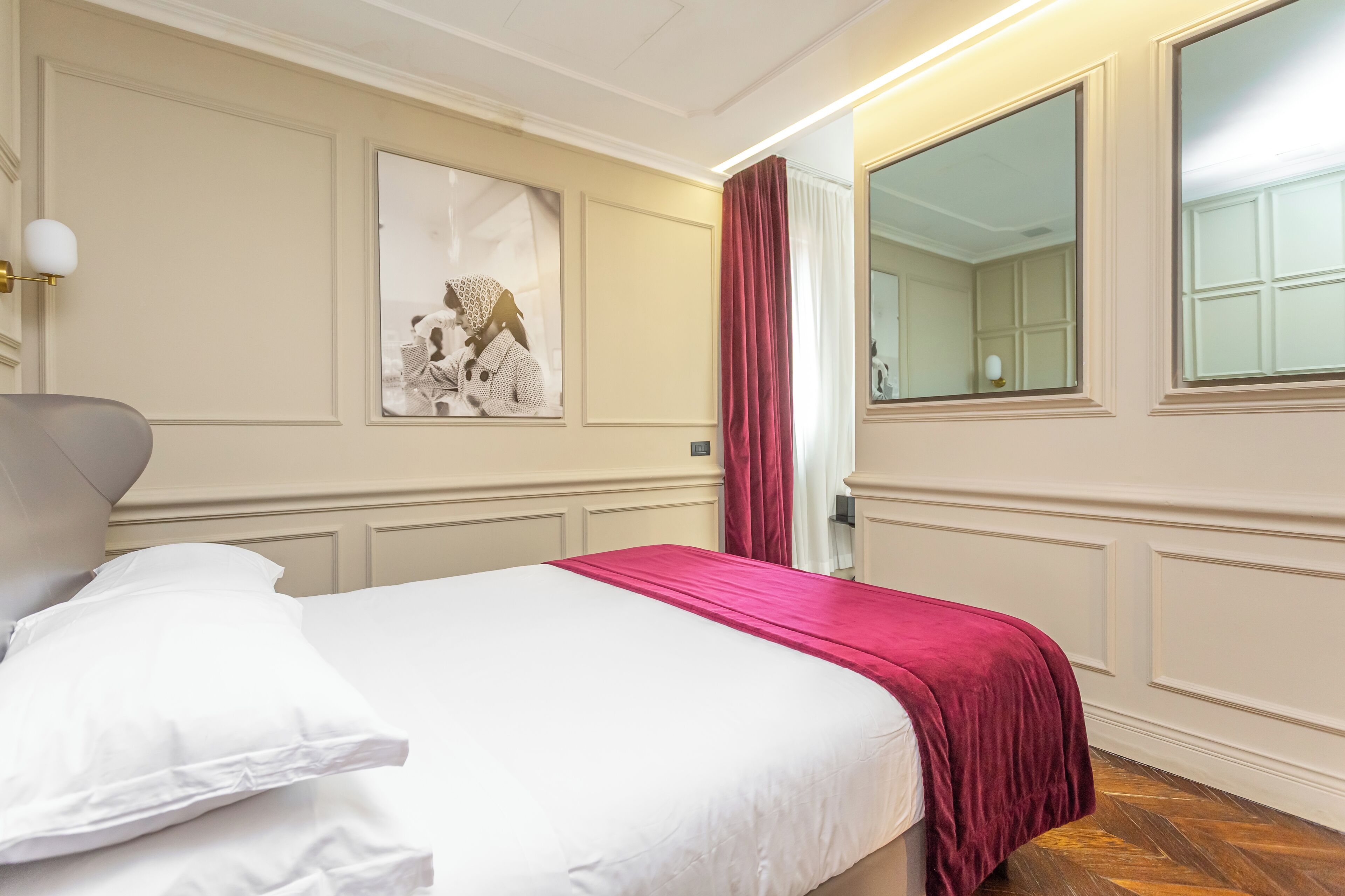 A hotel room with a bed, a mirror, a wall-mounted light, and a framed picture.