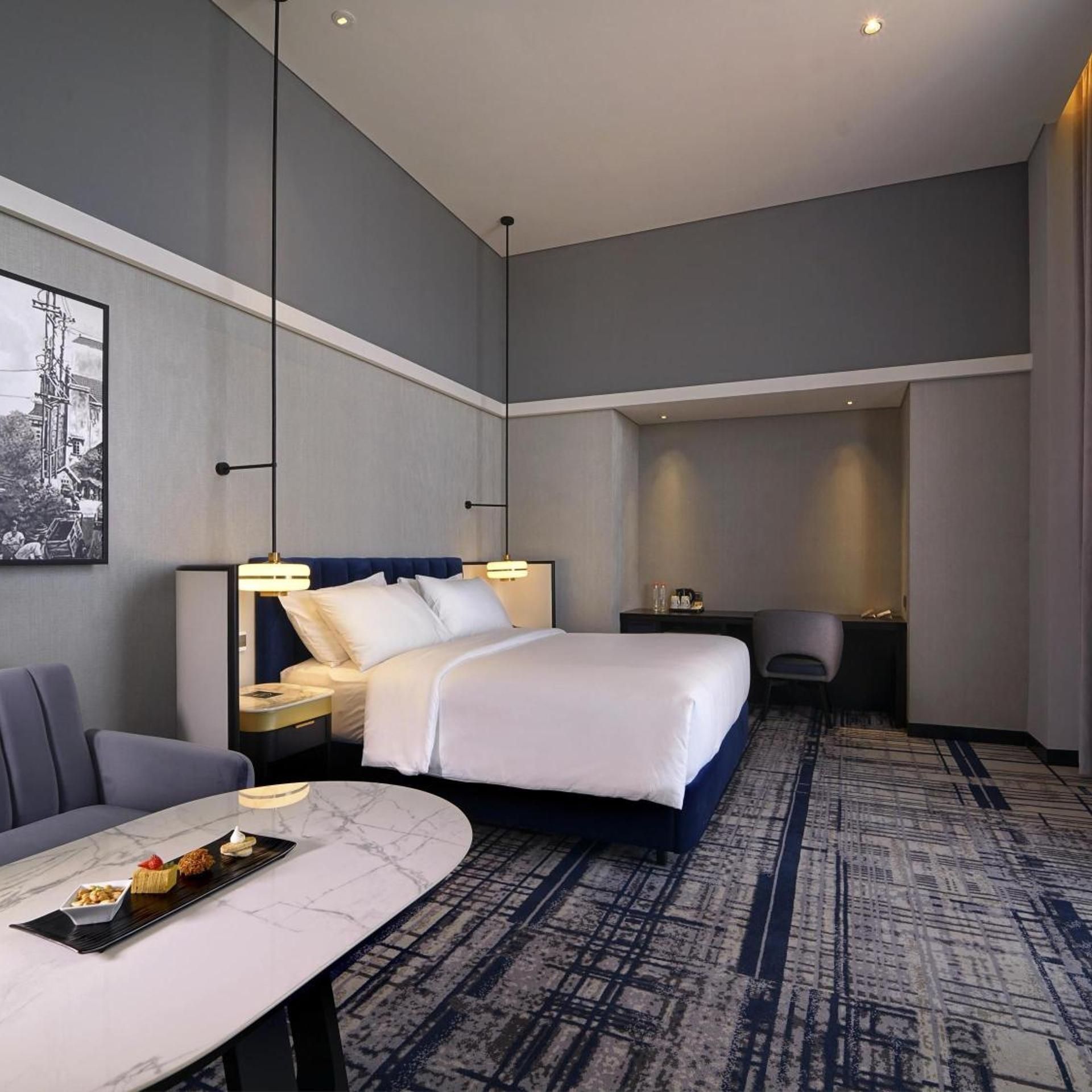 A hotel room with a bed, a desk, a chair, a dining table with a tray of food, and a framed picture on the wall.