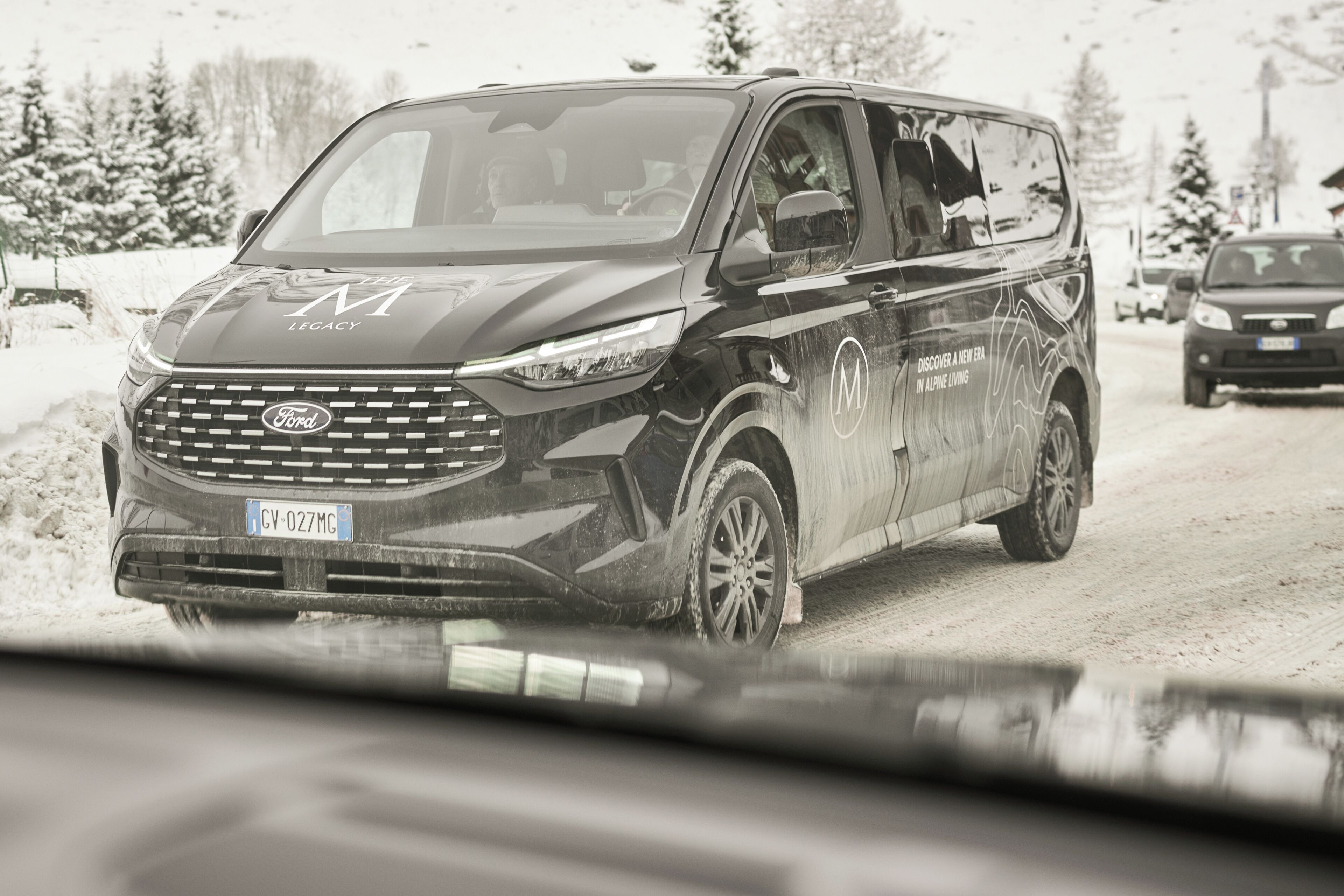 A black Ford Transit van with a license plate CV-027MC, driving on a snowy road.