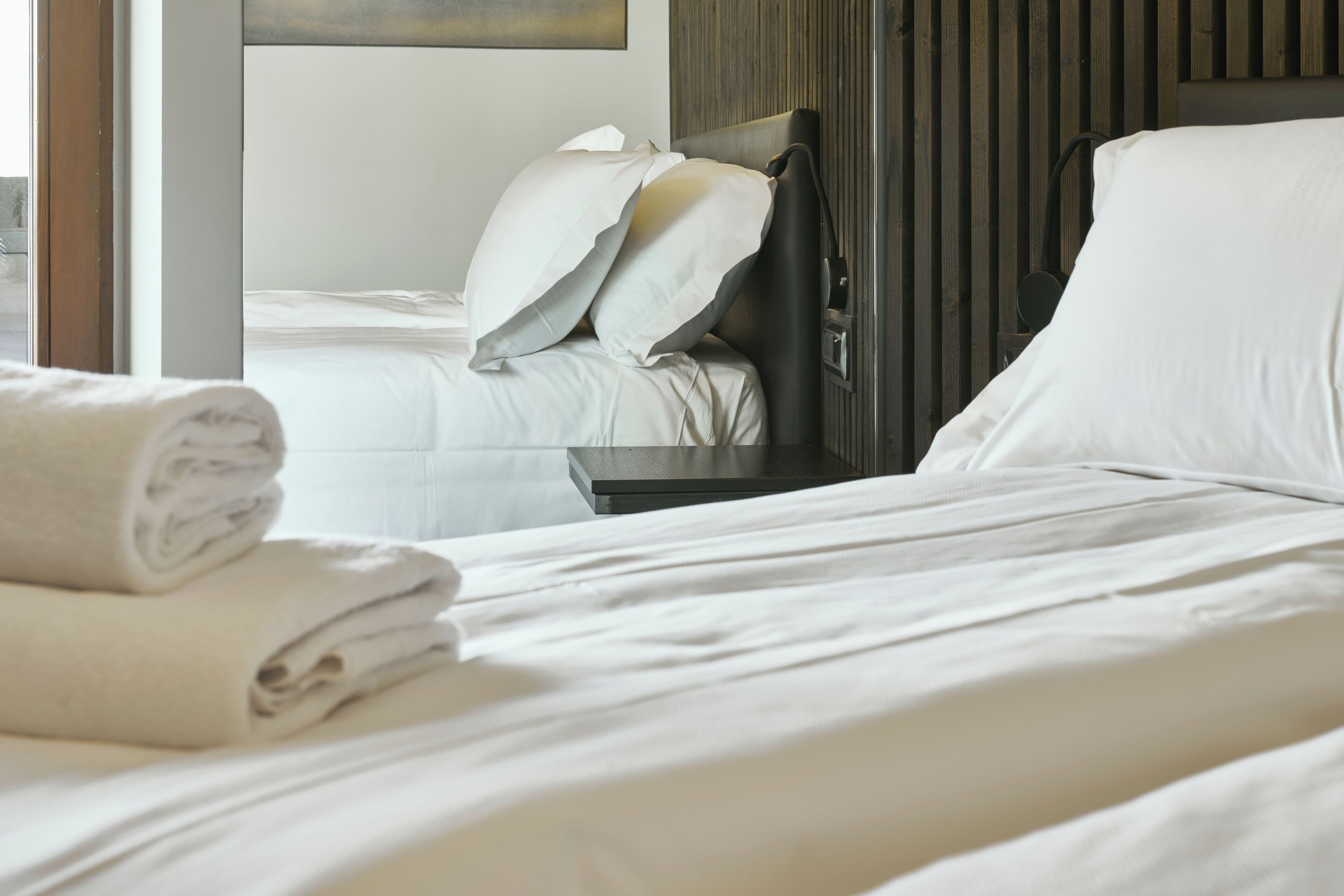 Une chambre d’hôtel avec un lit bien fait, une table de chevet et une fenêtre donnant sur une autre chambre.