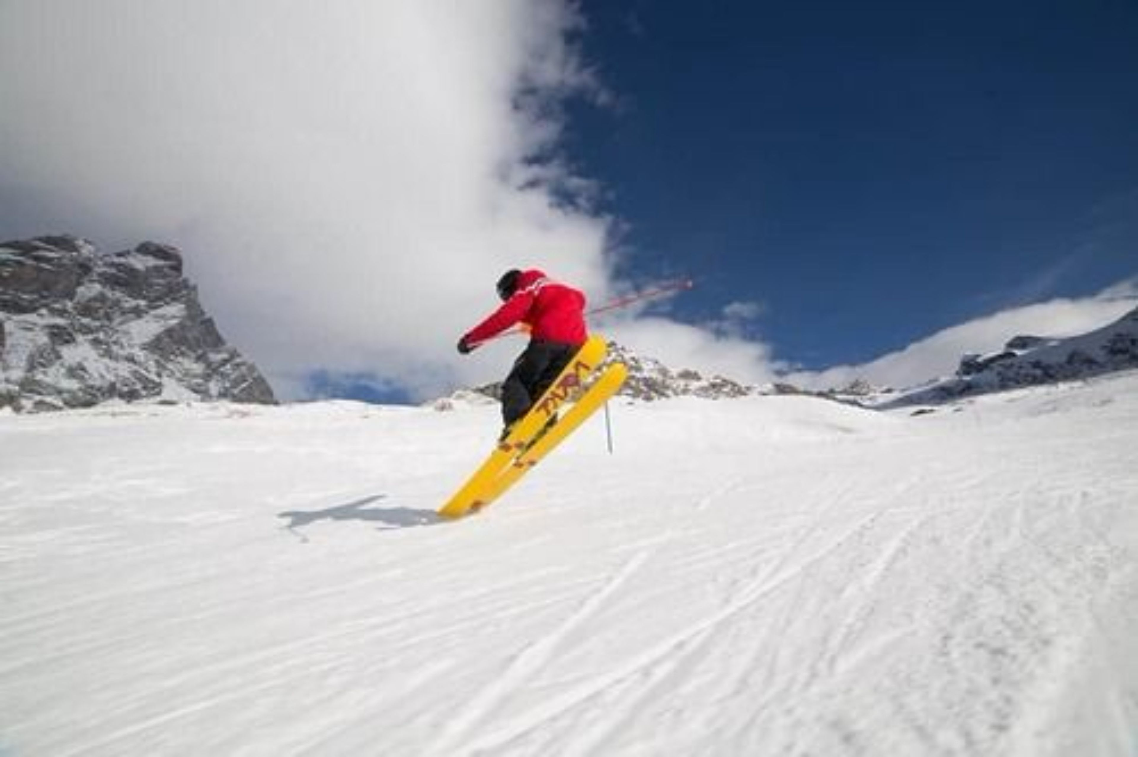 Un snowboarder con chaqueta roja realizando un salto en una ladera nevada con montañas al fondo.