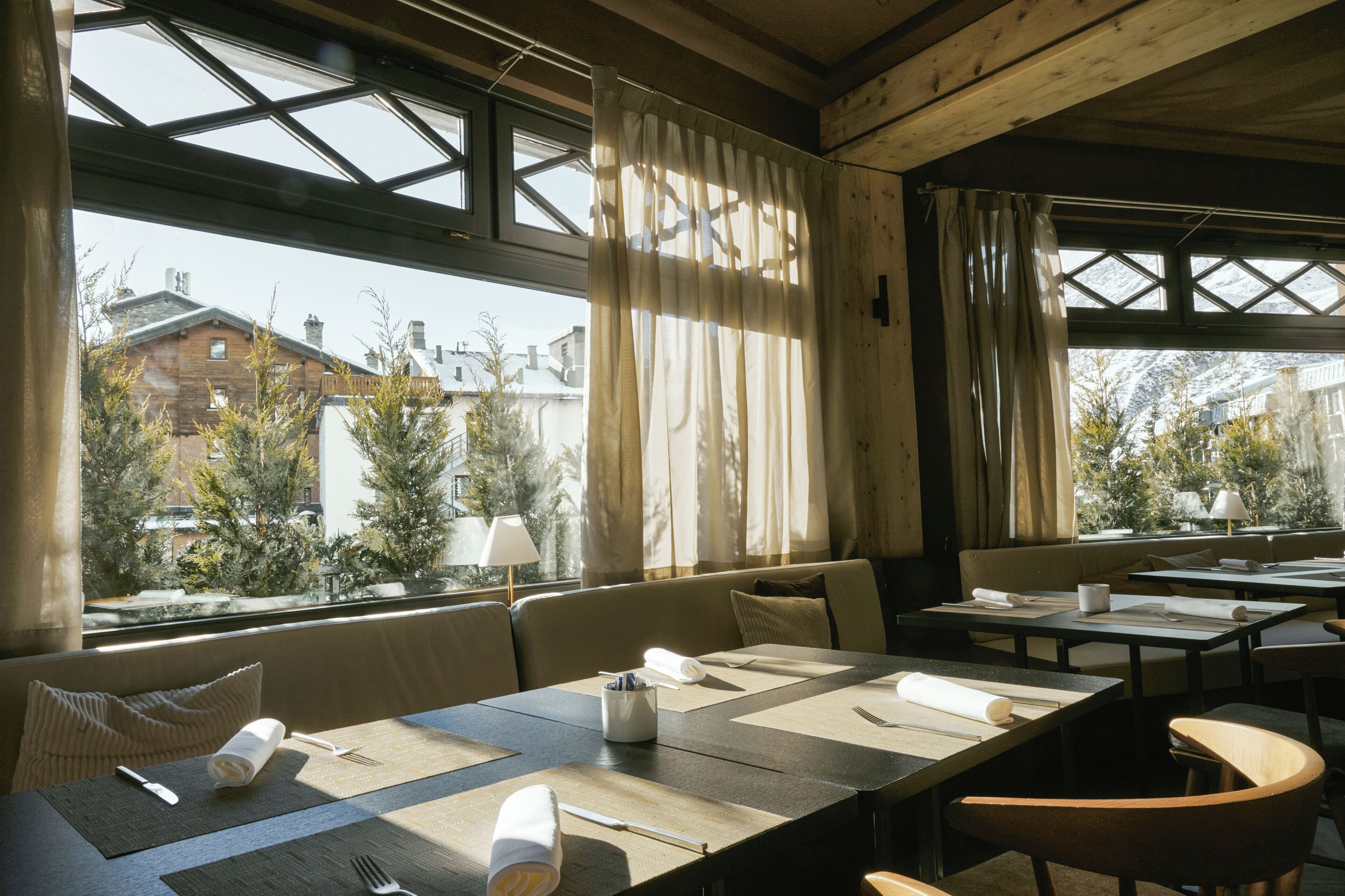 A dining area with tables set for two, large windows with curtains, and a view of buildings and trees outside.
