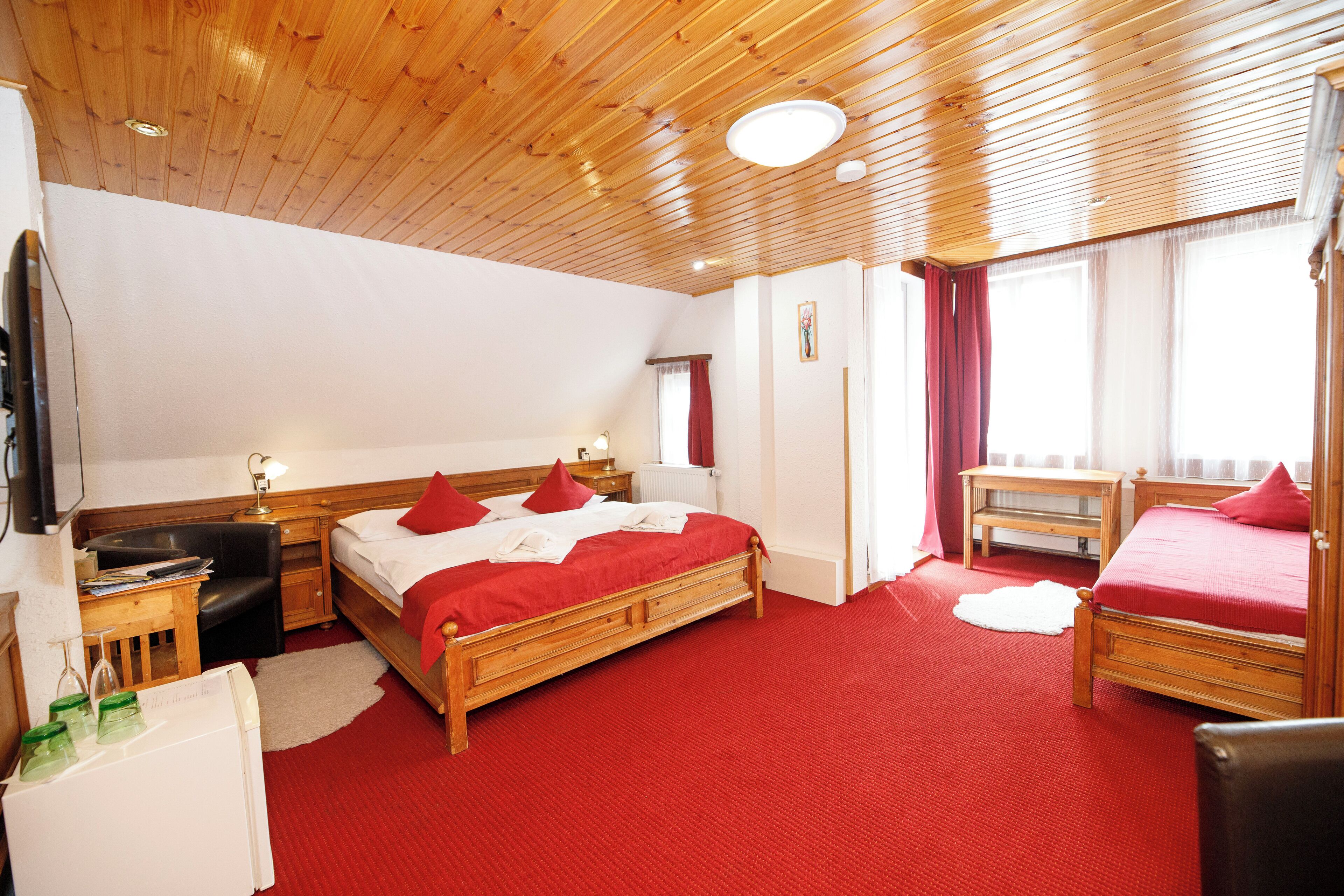A hotel room with a wooden ceiling, a large bed with red bedding, a desk with a lamp, and a small table with a chair.