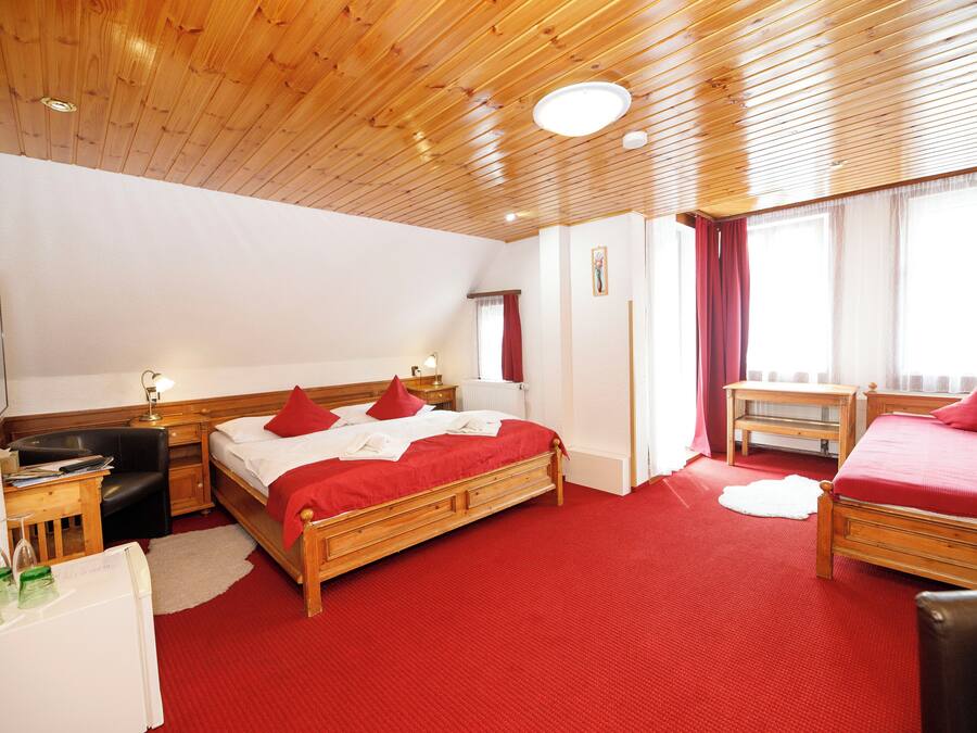 A hotel room with a wooden ceiling, a large bed with red bedding, a desk with a lamp, and a small table with a chair.