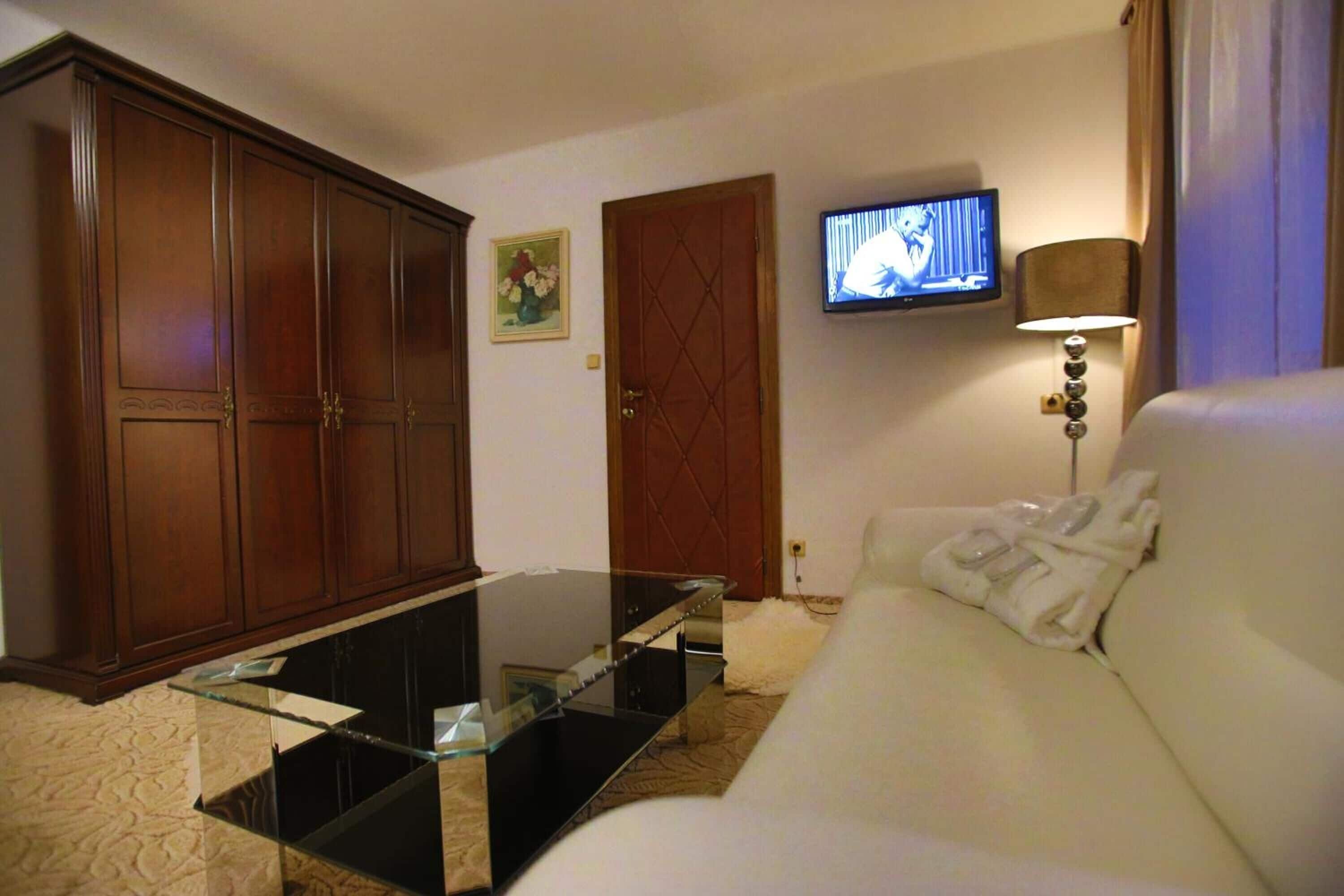 A living room with a glass coffee table, a white sofa, a wooden wardrobe, a television, and a lamp.