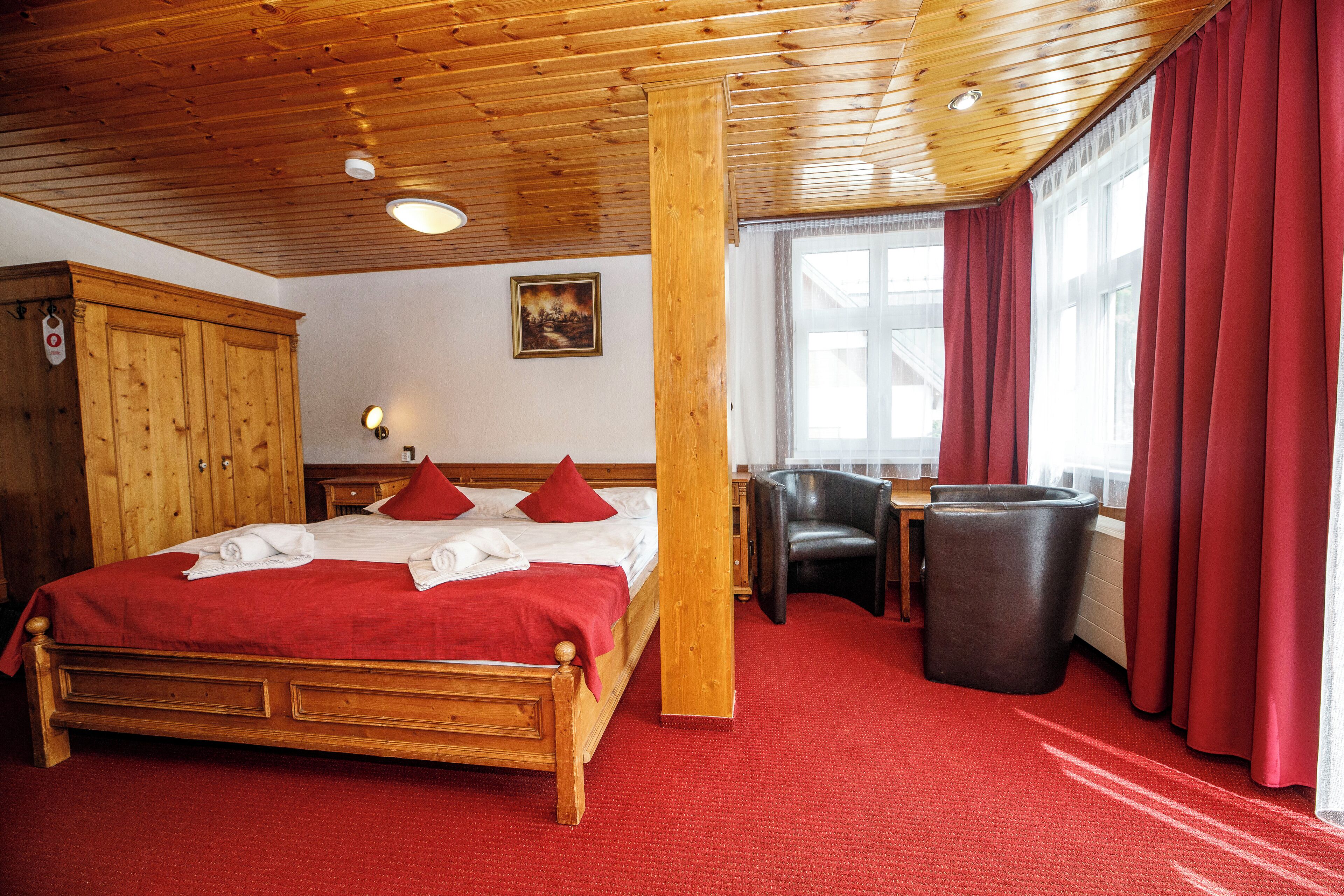 A wooden-framed bed with red bedding, a wooden wardrobe, a chair, and a trash bin in a room with red carpet and wooden ceiling beams.