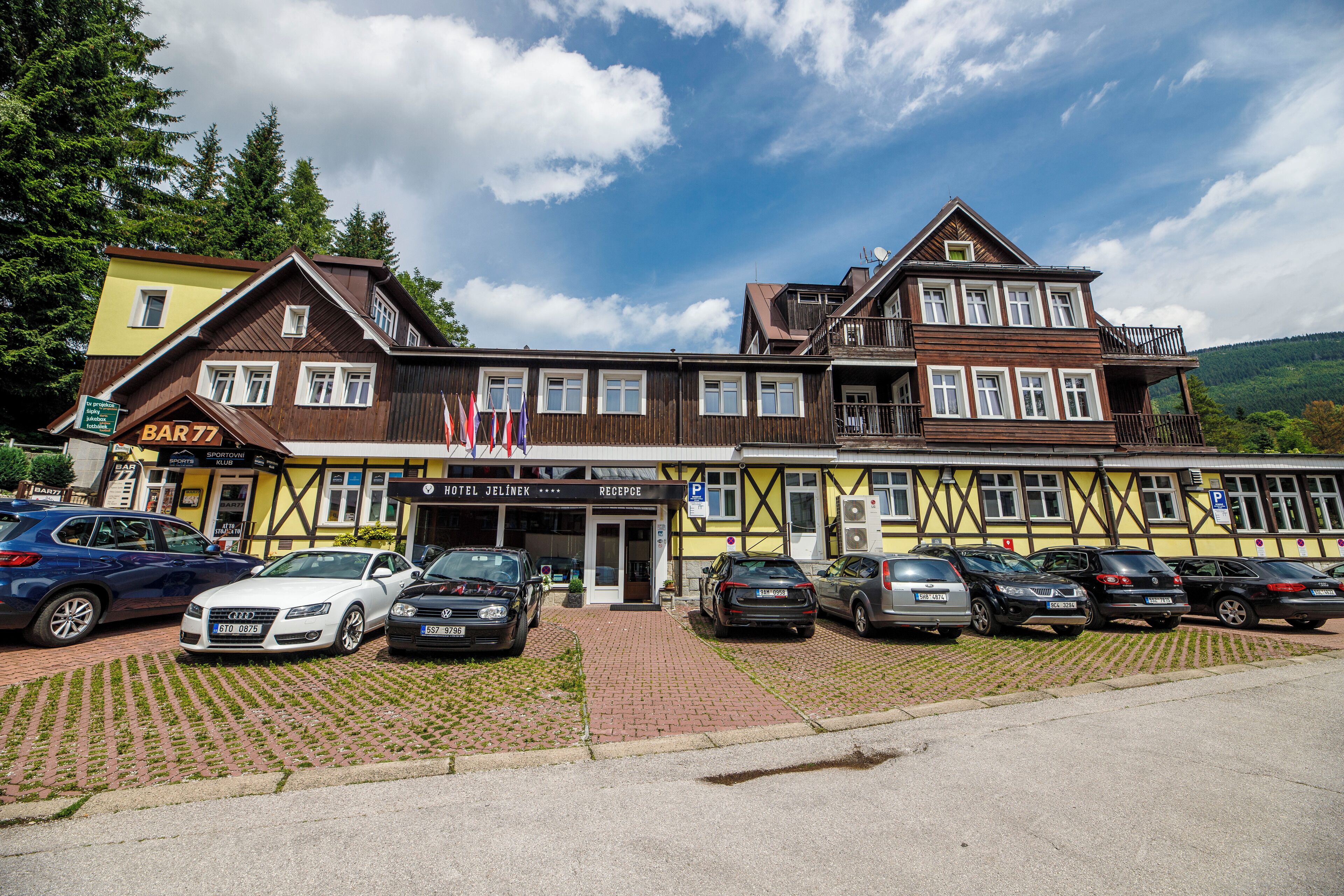 A traditional building with a sign for 'BAR 77' and 'HOTEL JELINEK'. There are multiple cars parked in front, and the building has a balcony with flags.
