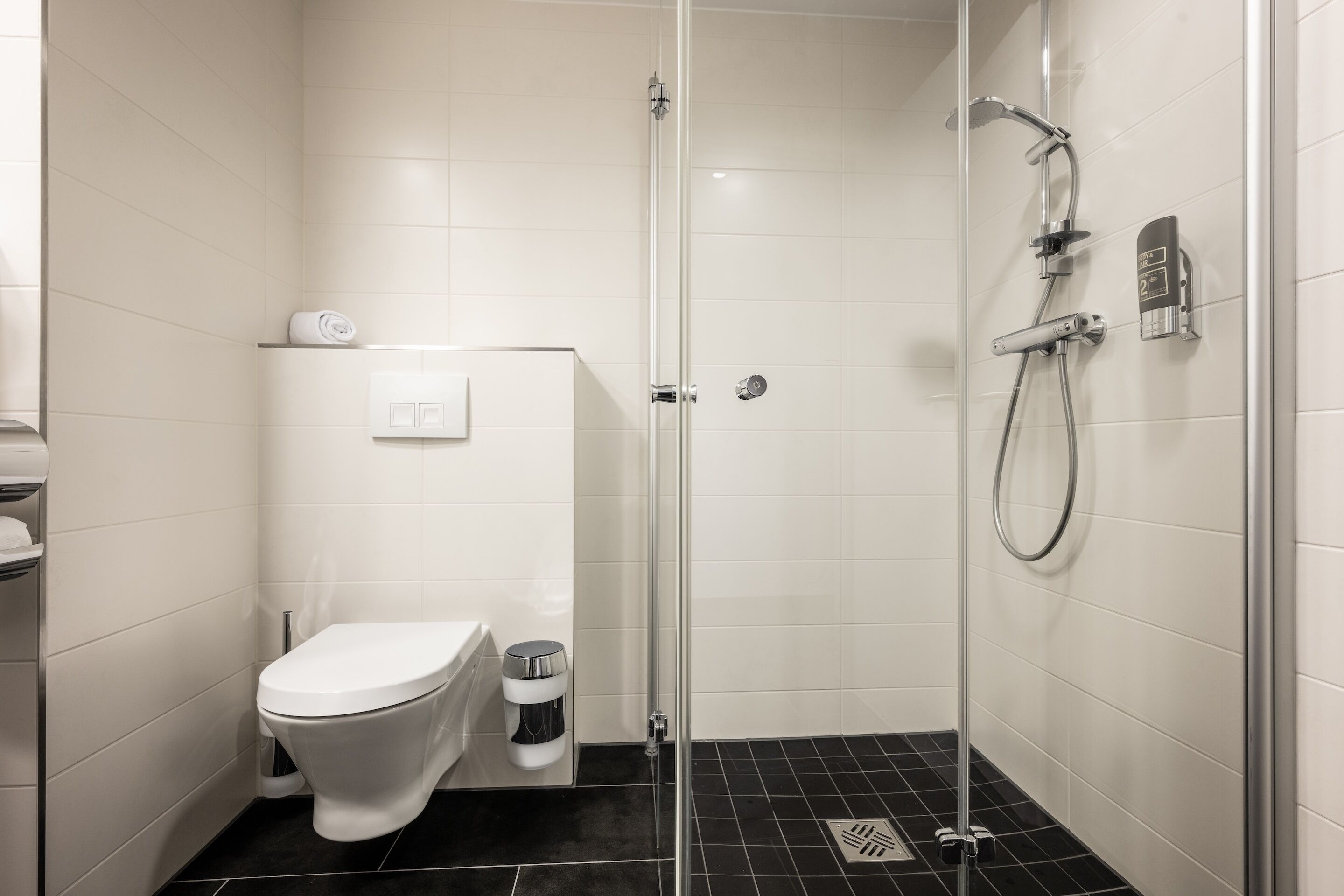 Un bany modern amb plat de dutxa envoltat de vidre, un vàter i un dispensador de tovalloles.