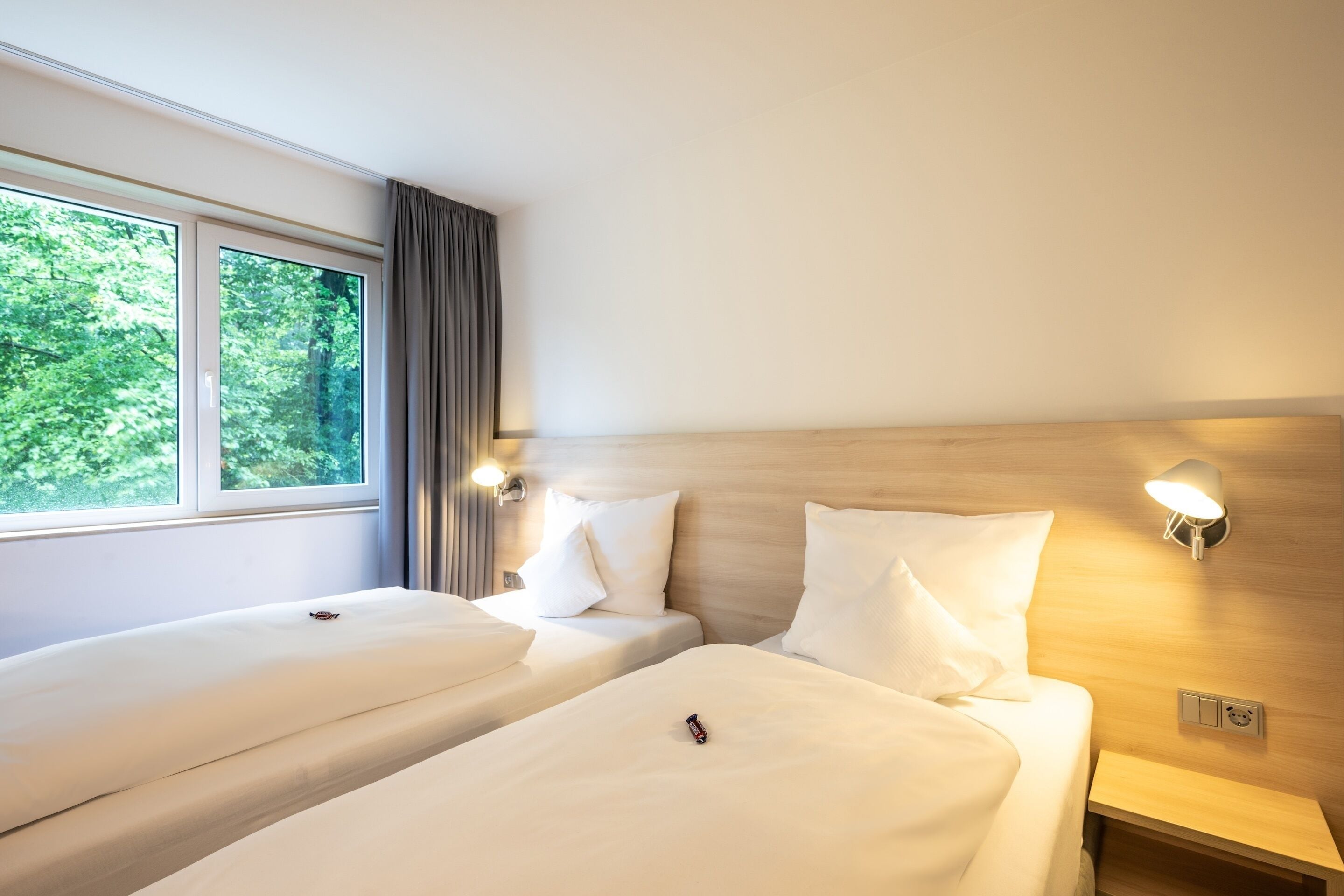Une chambre d’hôtel avec deux lits, une grande fenêtre avec des rideaux, un lampadaire fixé au mur et une tête de lit en bois.