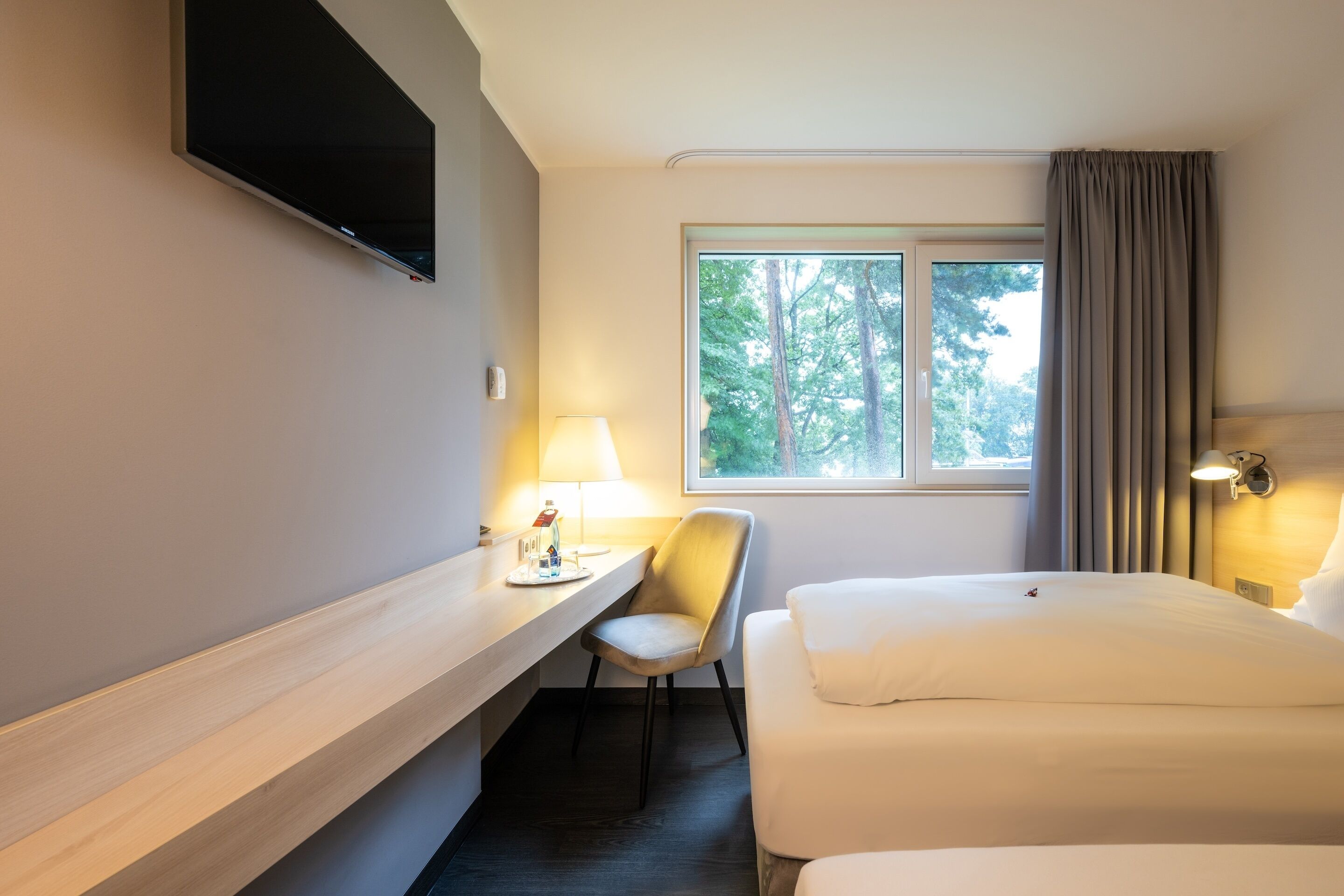Une chambre d’hôtel équipée d’un lit, d’un bureau, d’une chaise, d’un téléviseur fixé au mur et d’une fenêtre avec des rideaux.