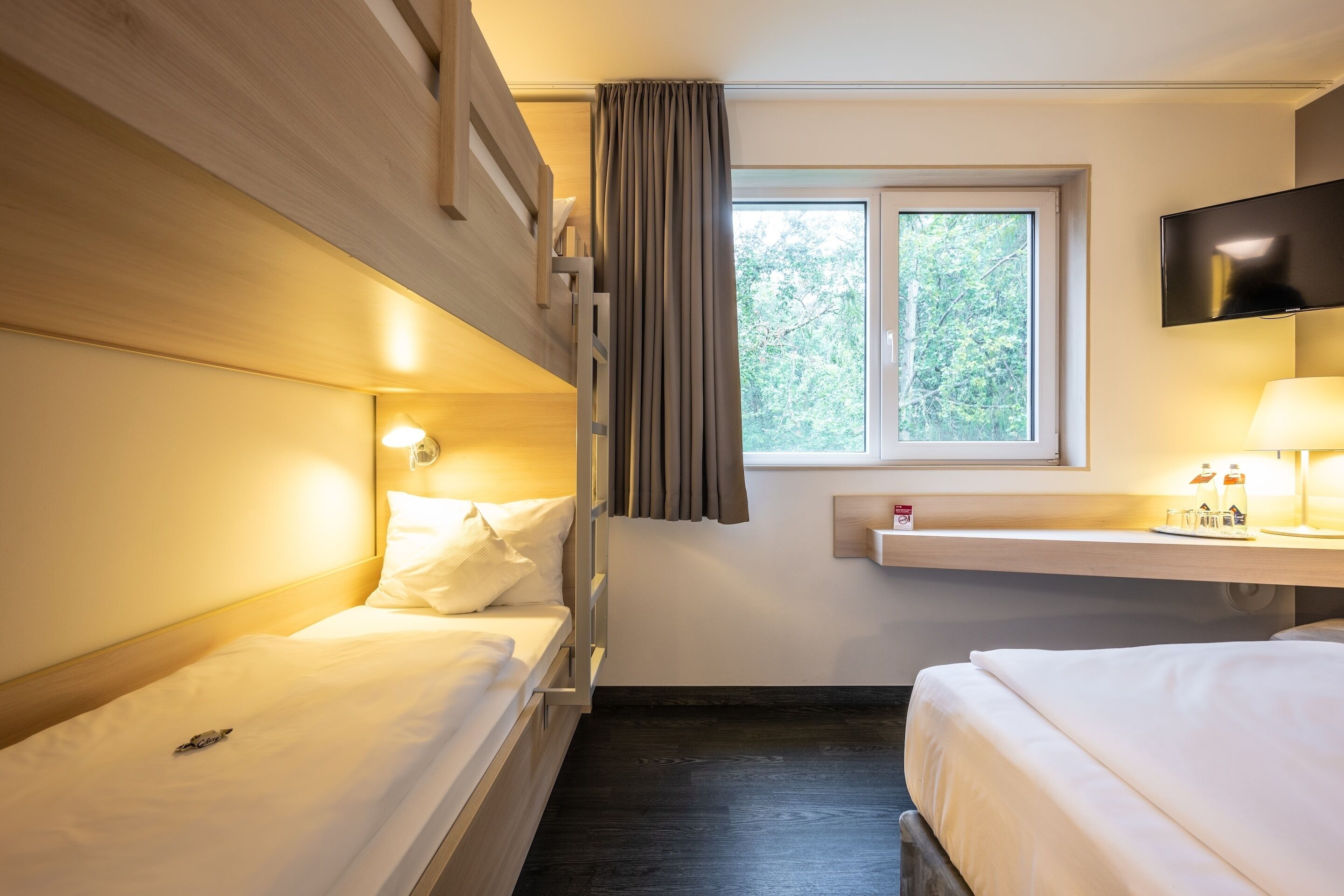 A hotel room with two beds, a TV mounted on the wall, a window with curtains, and a bedside table with a lamp.