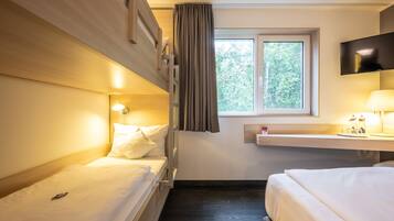 A hotel room with two beds, a TV mounted on the wall, a window with curtains, and a bedside table with a lamp.