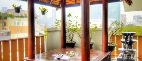 A thatched-roof gazebo with a table and chairs, potted plants, and a water feature on a balcony with a city view.