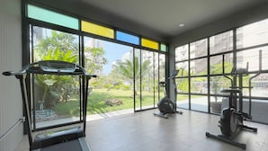 A gym with a treadmill, an elliptical, and a weight machine, large windows with a view of greenery and buildings. - Sabai Inn (Pattaya)