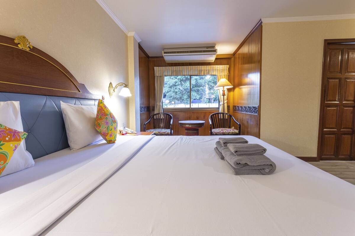 Quarto de hotel com uma cama grande, cabeceira de madeira, duas cadeiras, uma mesinha e uma janela com cortinas.