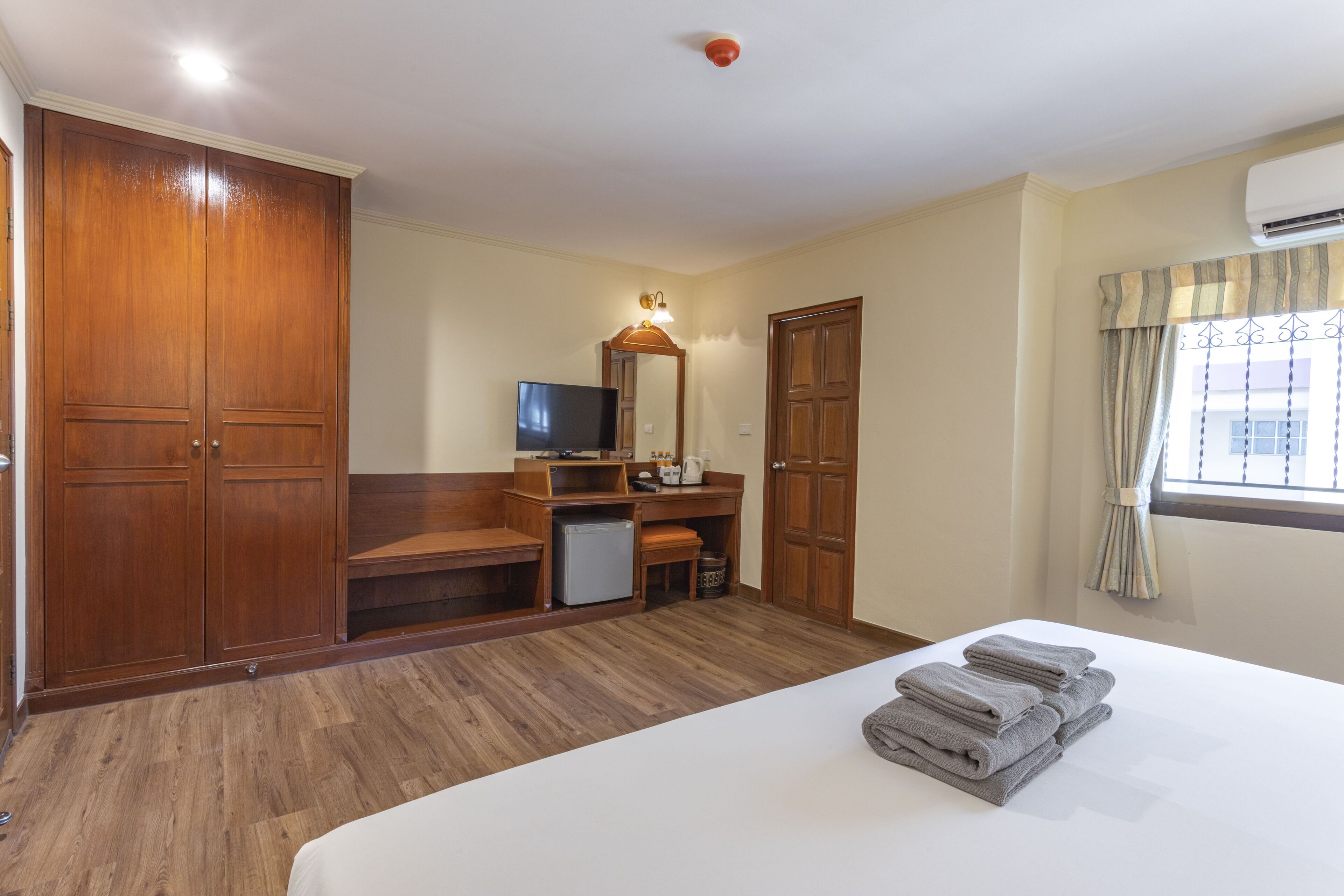 Une chambre d’hôtel dotée d’une grande armoire, d’un bureau avec un miroir, d’un petit réfrigérateur et d’un lit bien fait, agrémenté de serviettes.
