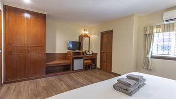 A hotel room with a large wardrobe, a desk with a mirror, a small refrigerator, and a neatly made bed with towels.