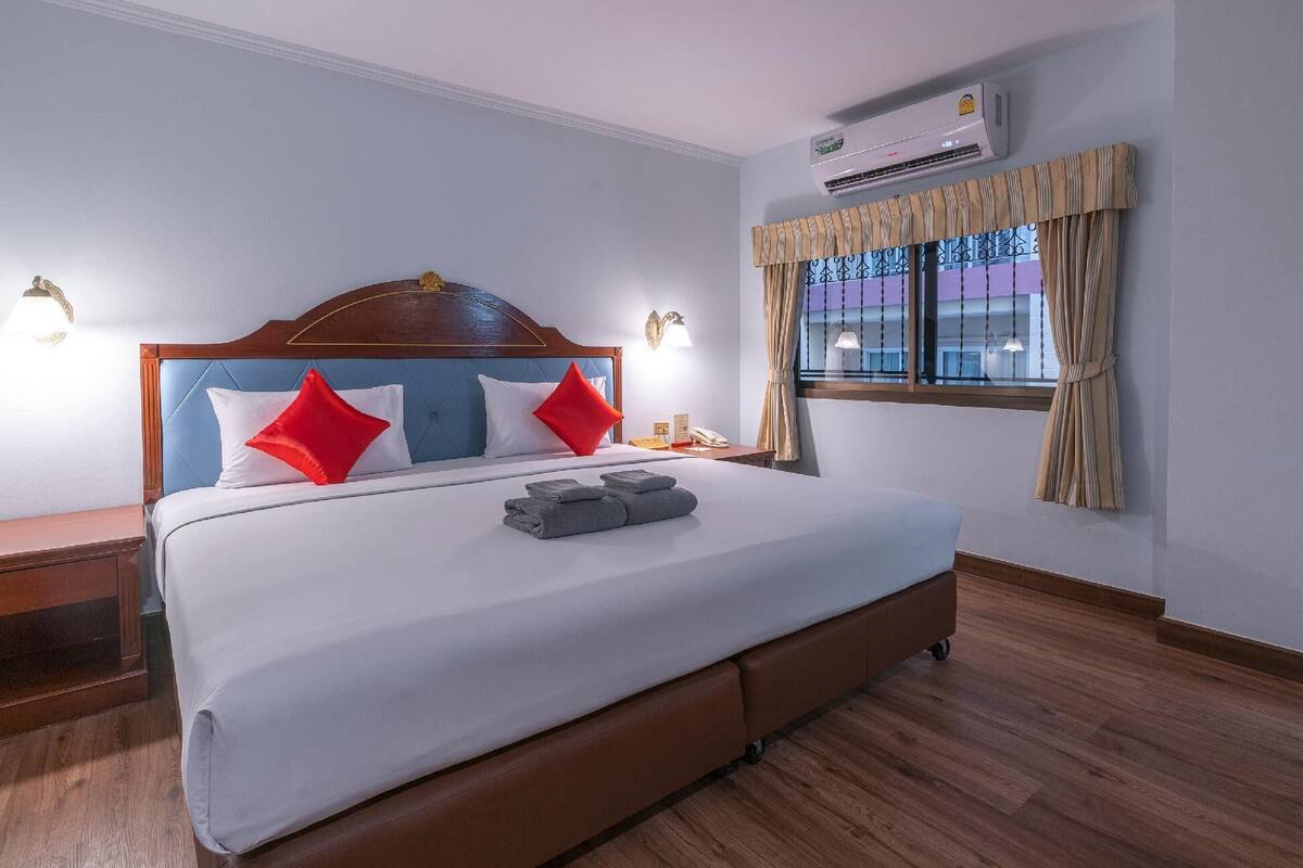 Quarto de hotel com uma cama grande, cabeceira de madeira, duas luminárias de cabeceira, janela com cortinas, ar condicionado e piso de madeira.