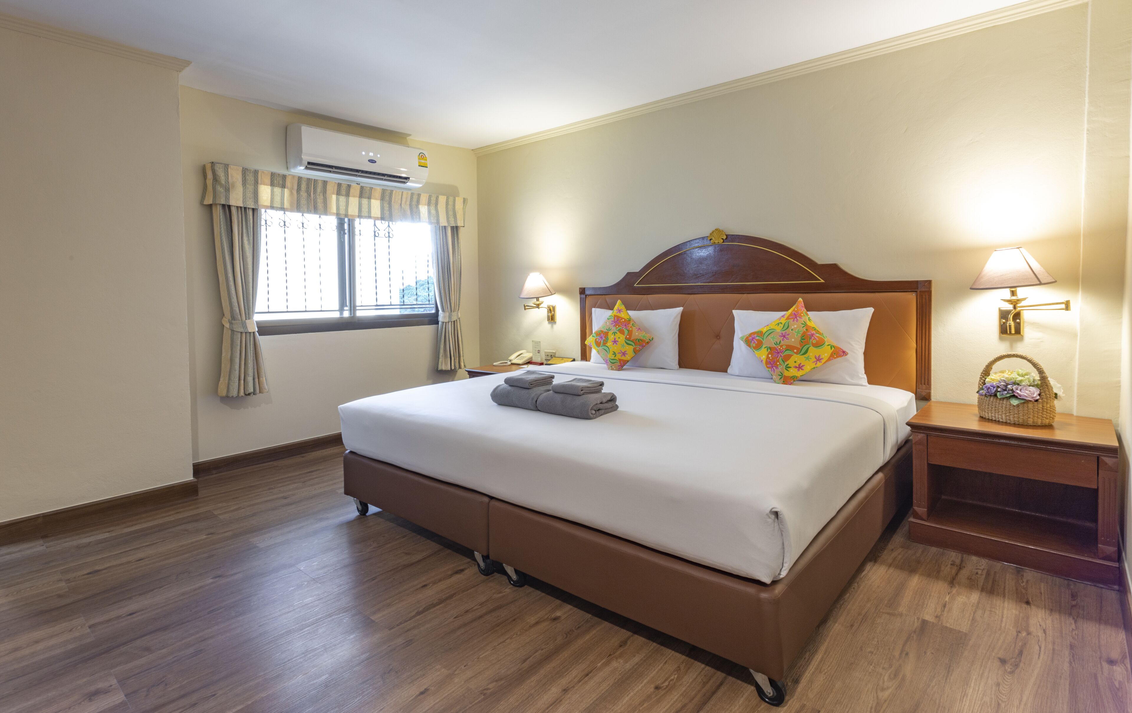 Una habitación de hotel con una cama grande, cabecera de madera, dos lámparas de noche, una ventana con cortinas y un equipo de aire acondicionado.