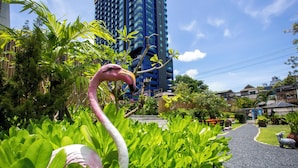 A garden with a pink flamingo statue, lush greenery, and a pathway leading to a building with a blue facade. - Sabai Inn (Pattaya)