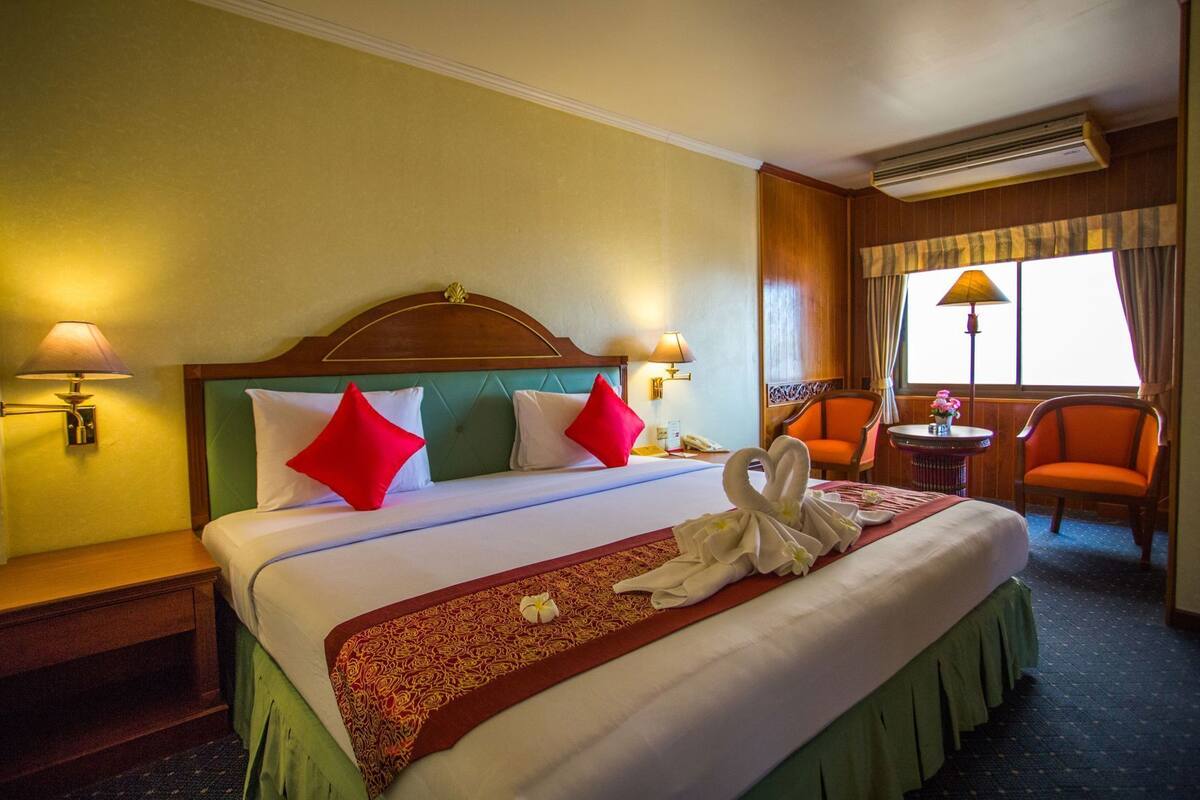 Quarto de hotel com uma cama grande, duas luminárias laterais, uma cabeceira de madeira, uma pequena mesa com abajur, uma cadeira laranja e uma janela com cortinas.