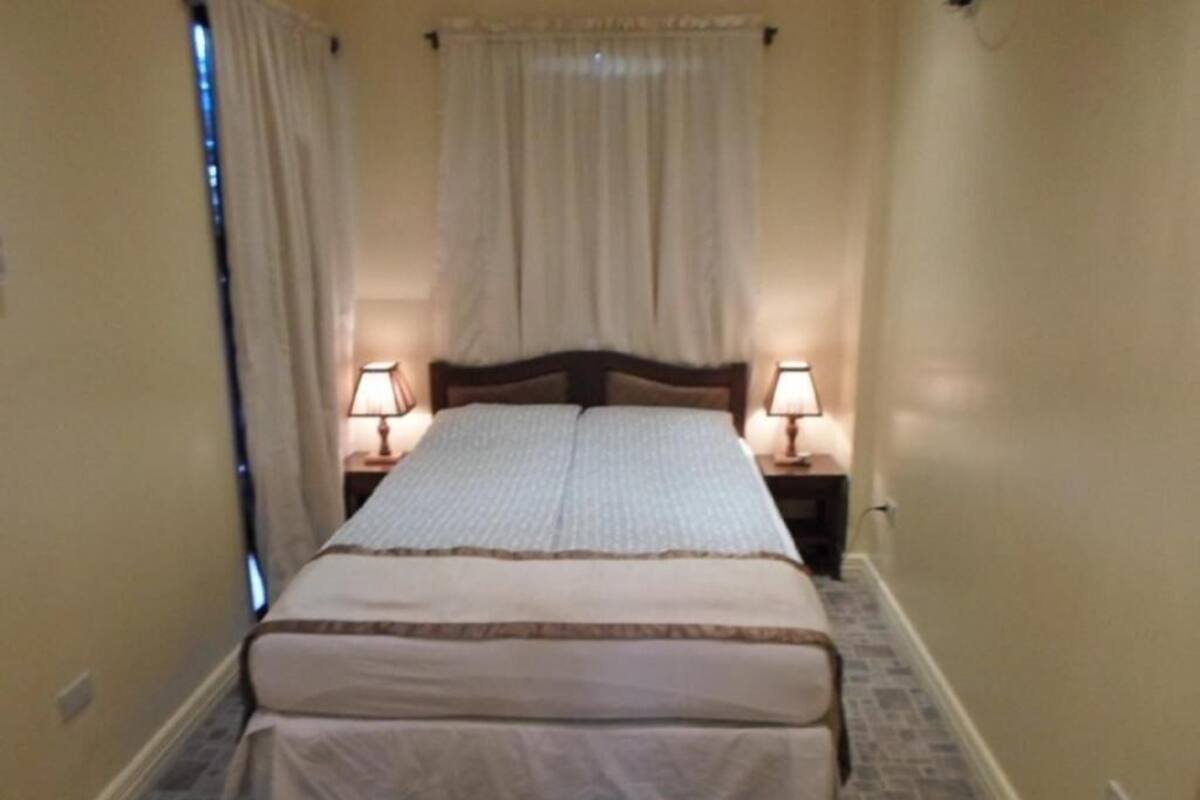 Un dormitorio con una cama doble, dos mesitas de noche con lámparas, una ventana con cortinas y suelo de baldosas.