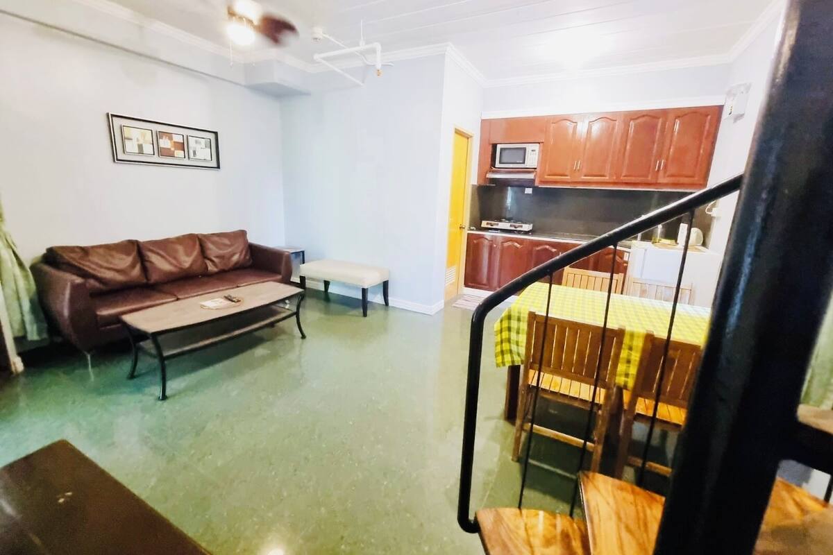 Una sala de estar con un sofá de cuero marrón, una mesita y un banco. Se ve una escalera que conduce a un comedor con una mesa y sillas de madera. Al fondo, una cocina con armarios de madera y un microondas.