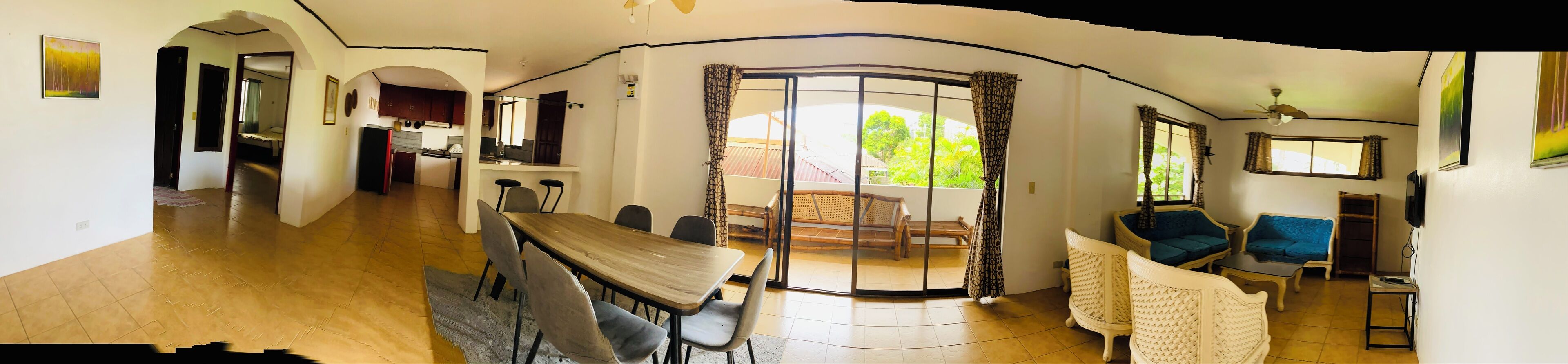 A spacious living area with a dining table and chairs, a sofa, and a balcony with a view of greenery.