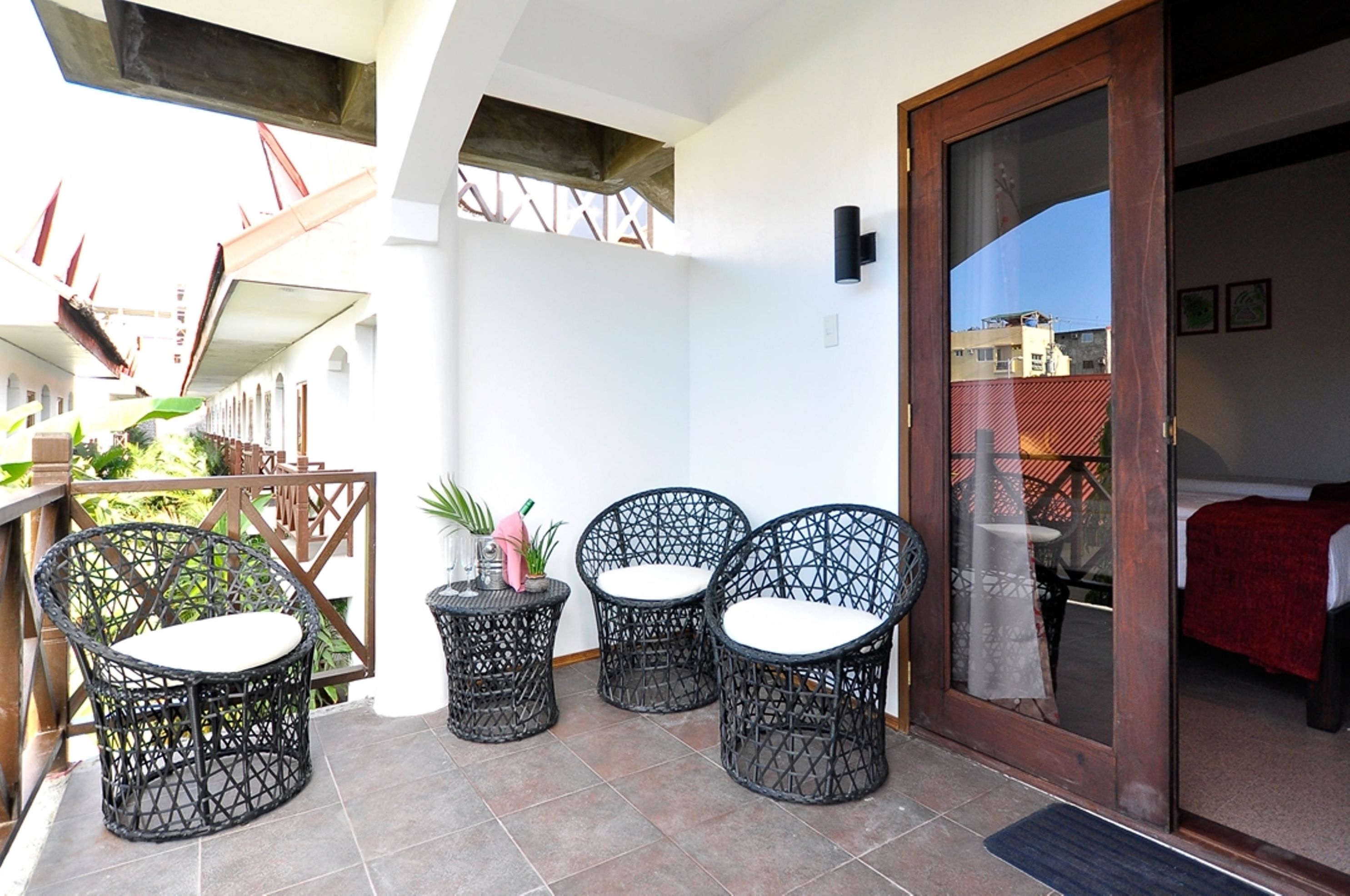 A balcony with wicker chairs and a small table, a sliding glass door leading to a bedroom, and a view of a building in the background.