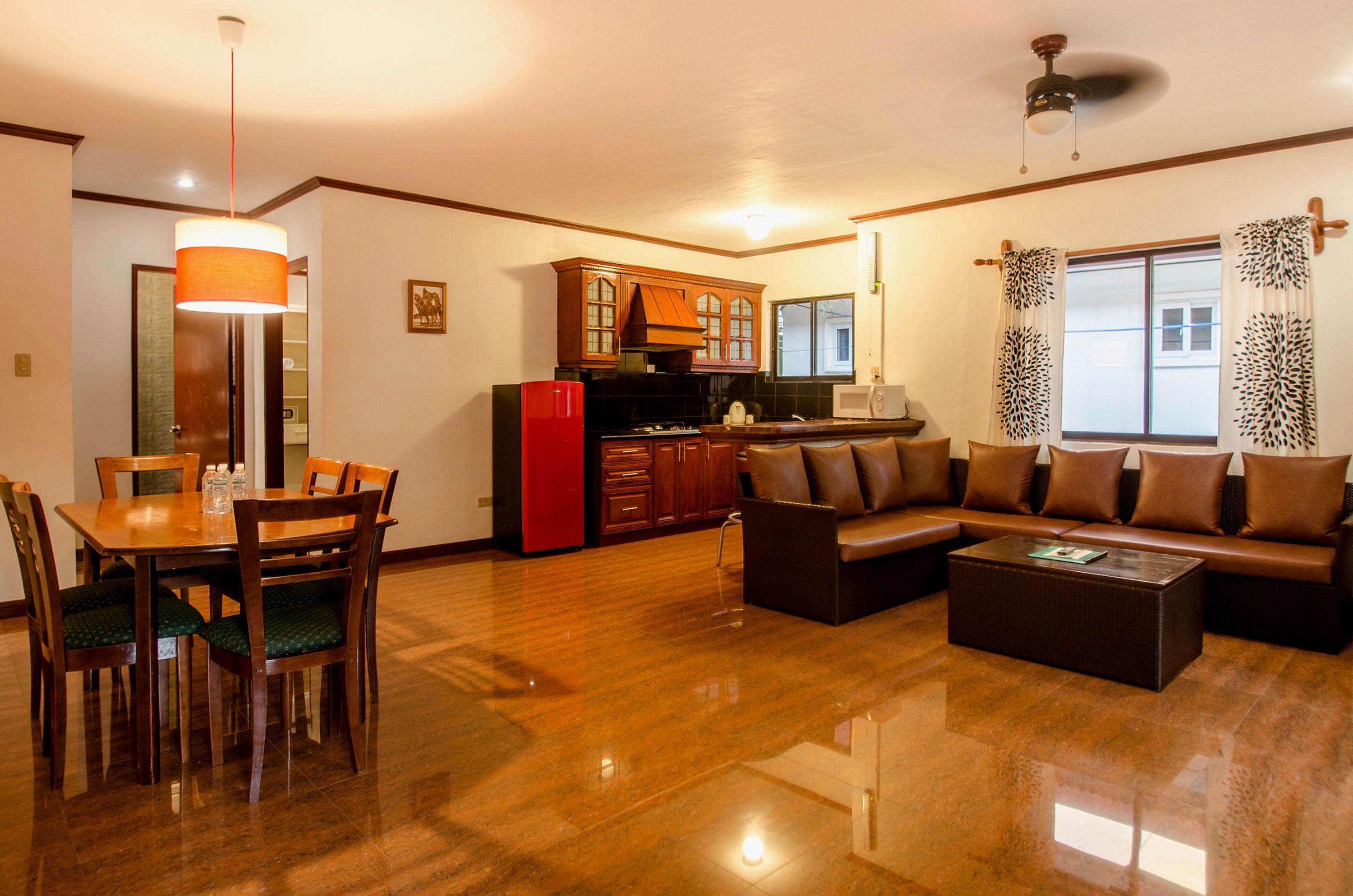 A spacious living area with a wooden dining table, chairs, a sofa, and a coffee table. A kitchen with wooden cabinets and a red refrigerator is visible in the background.