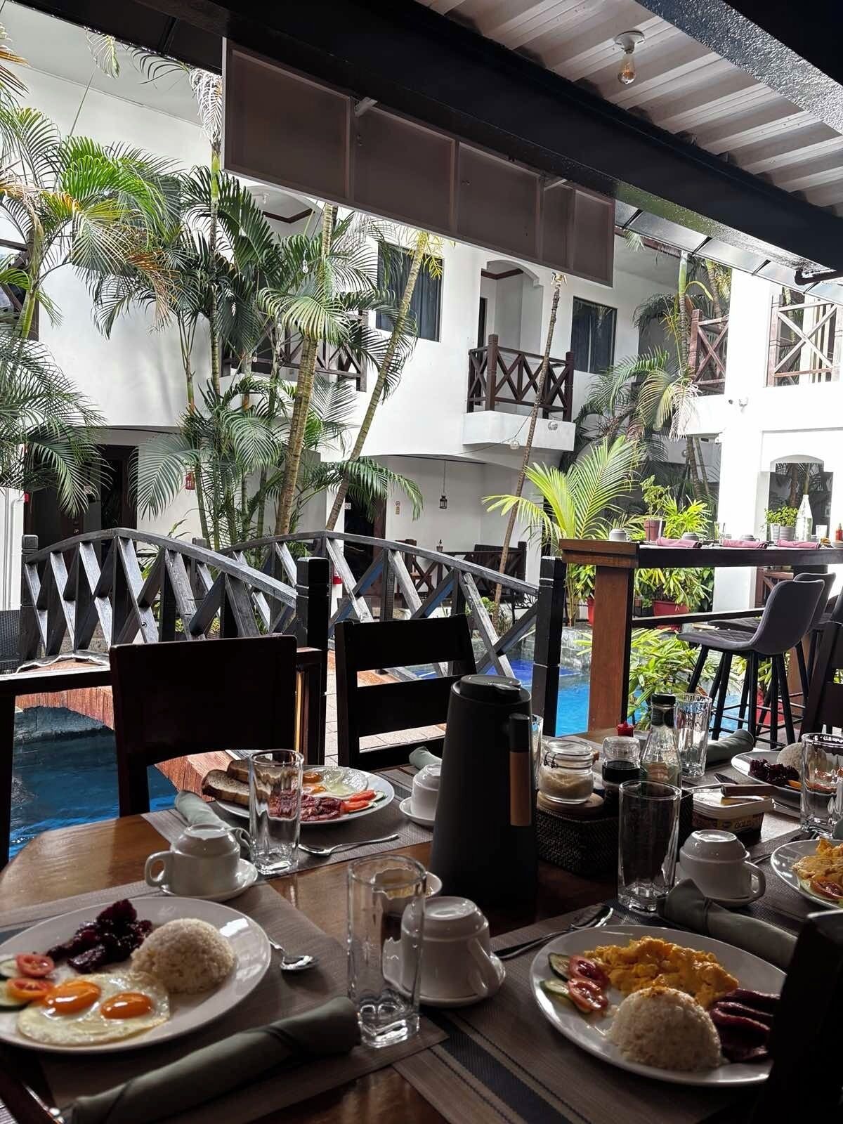 Un espacio al aire libre con mesas dispuestas para el desayuno, con vistas a una piscina y rodeado de exuberante vegetación.