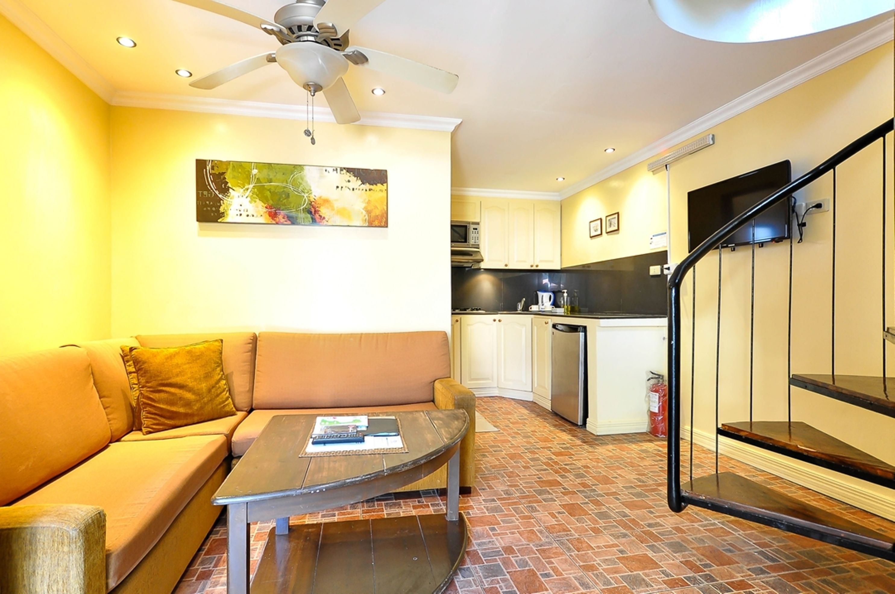 A living room with a sofa, coffee table, and a ceiling fan. Adjacent is a kitchen with cabinets and a microwave.