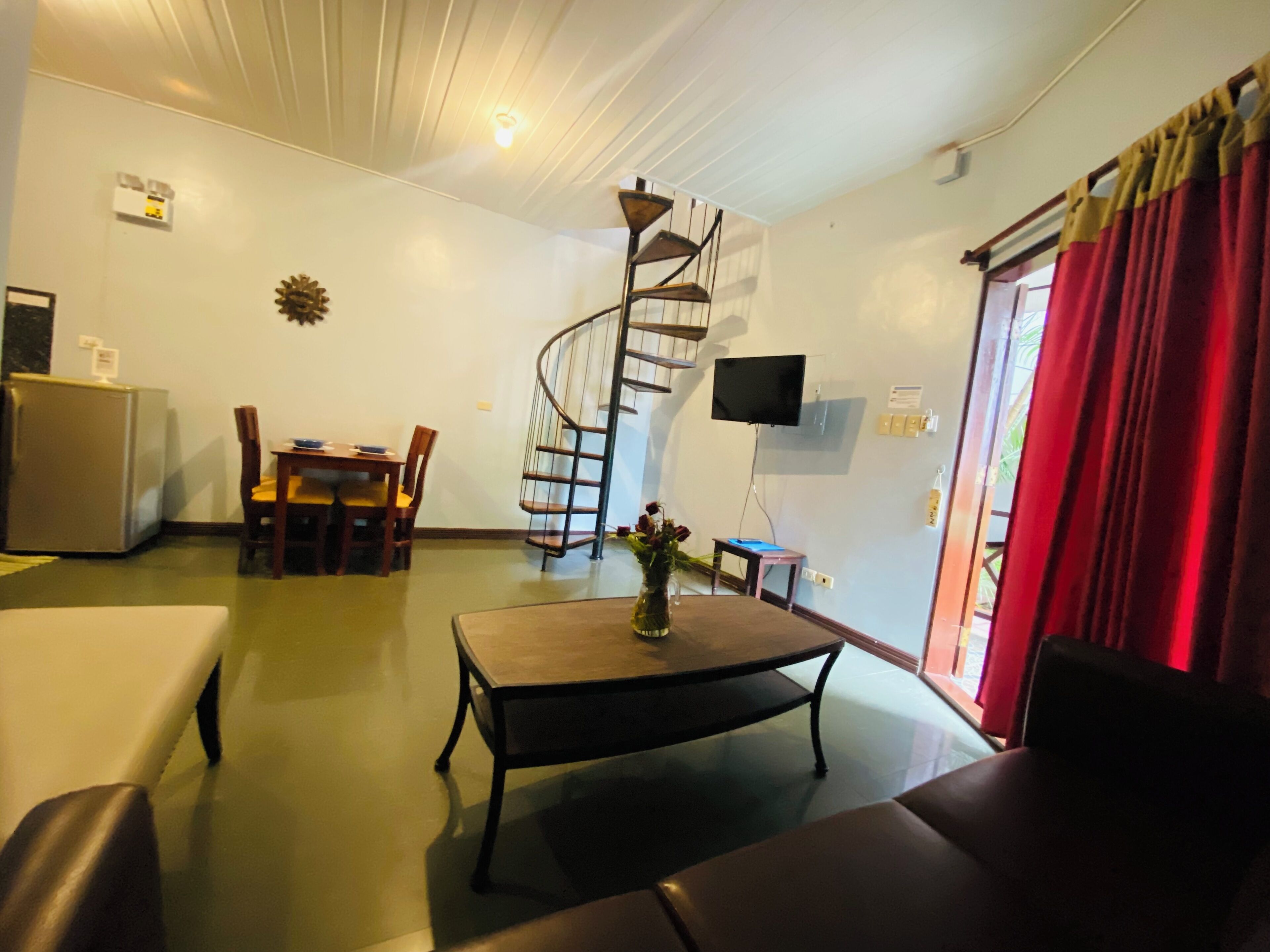 A living room with a spiral staircase, a dining area, a flat-screen TV, and a red curtain.