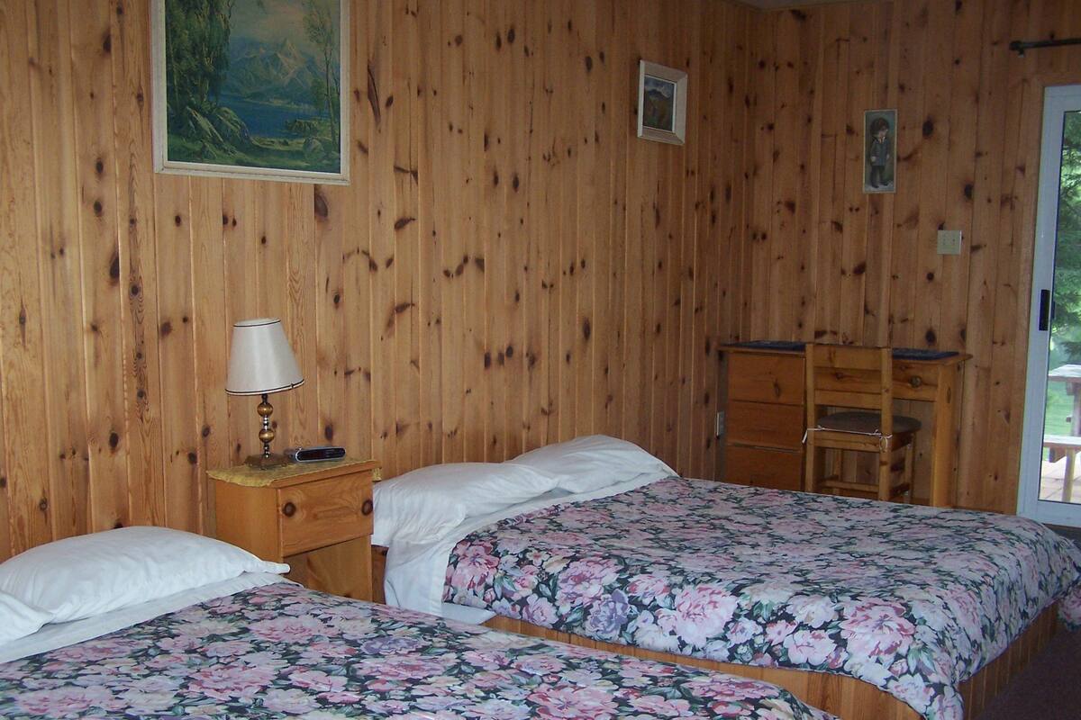 A room with wooden walls, two beds with floral bedding, a nightstand with a lamp, and a wooden dresser.