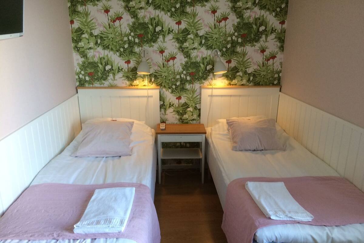 A room with two single beds, each with a white pillow and a pink blanket. There is a small wooden nightstand between the beds with a lamp on it. The wall behind the beds is decorated with a floral wallpaper.