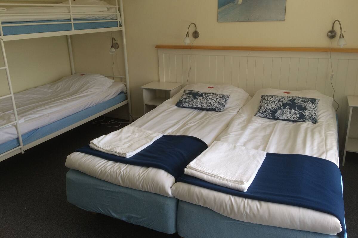A dormitory room with bunk beds, a single bed, a nightstand, a lamp, and a painting on the wall.