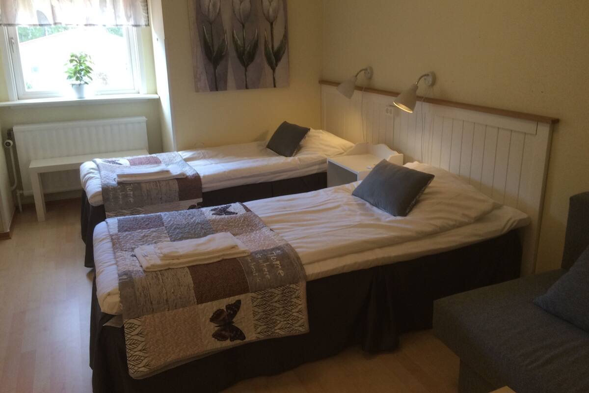 A room with two single beds, a bench, a radiator, and a window with curtains.