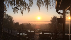 Une vue sur le lever du soleil depuis un balcon doté d’une balustrade en bois, donnant sur un paysage brumeux et une étendue d’eau.