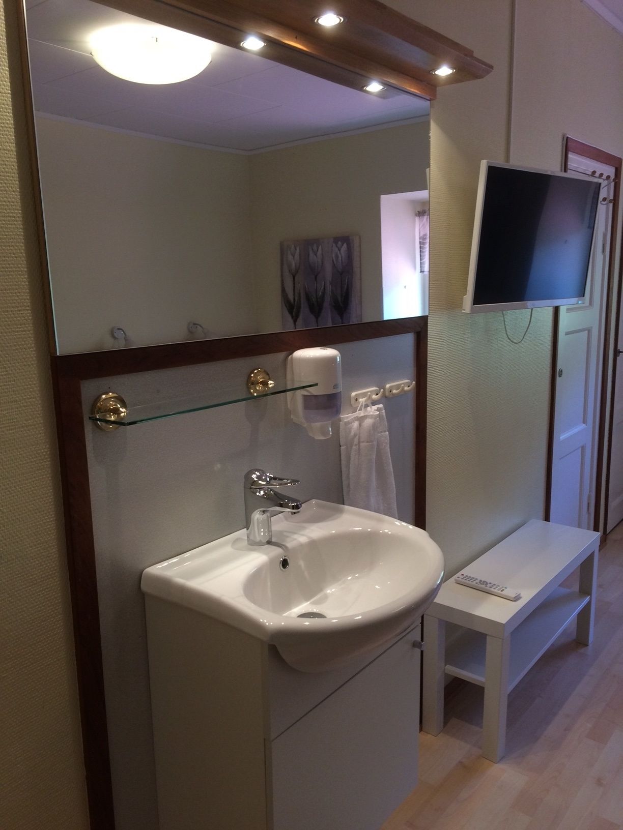 A bathroom with a white sink, a glass shelf with toiletries, a mirror, and a television mounted on the wall.