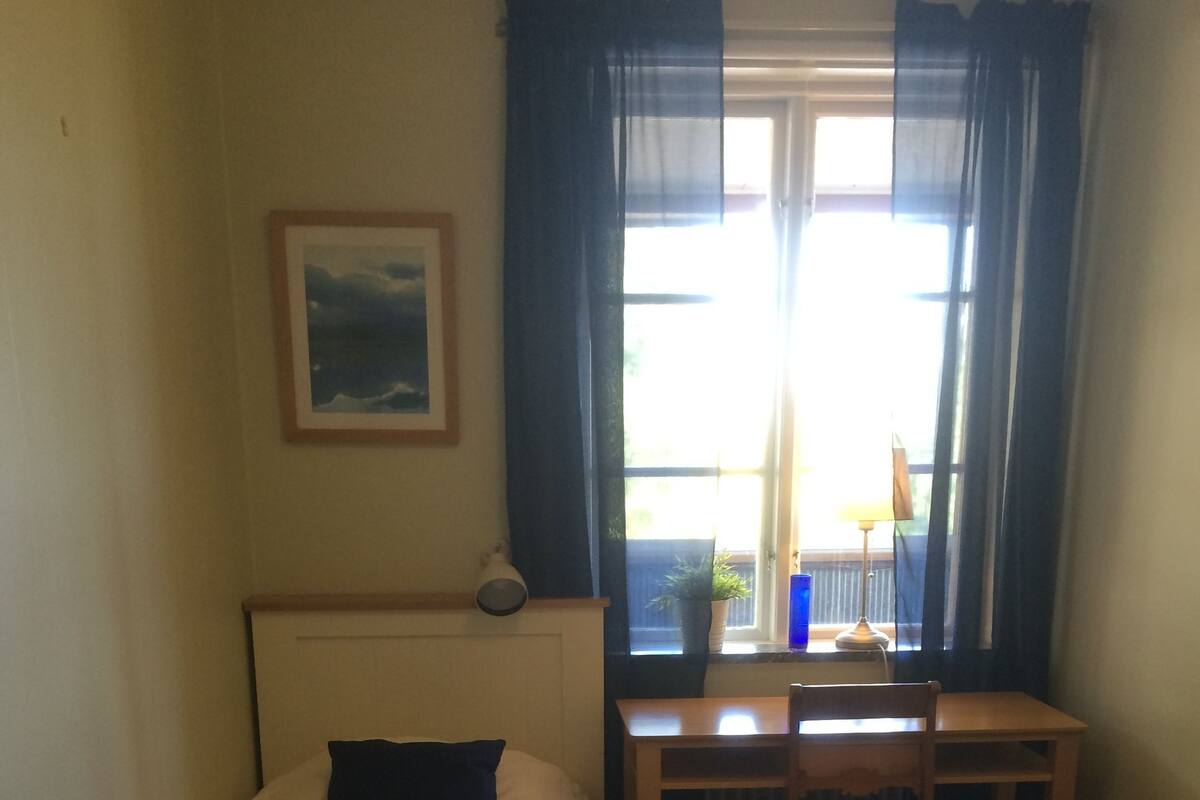 A room with a bed, a desk, and a chair. There is a window with curtains, a lamp on the desk, and a framed picture on the wall.