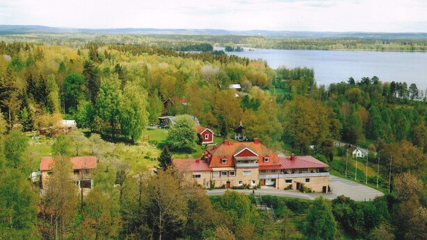 Un grand bâtiment à plusieurs étages, doté d’un toit rouge, entouré d’une forêt dense et avec un lac en arrière-plan.