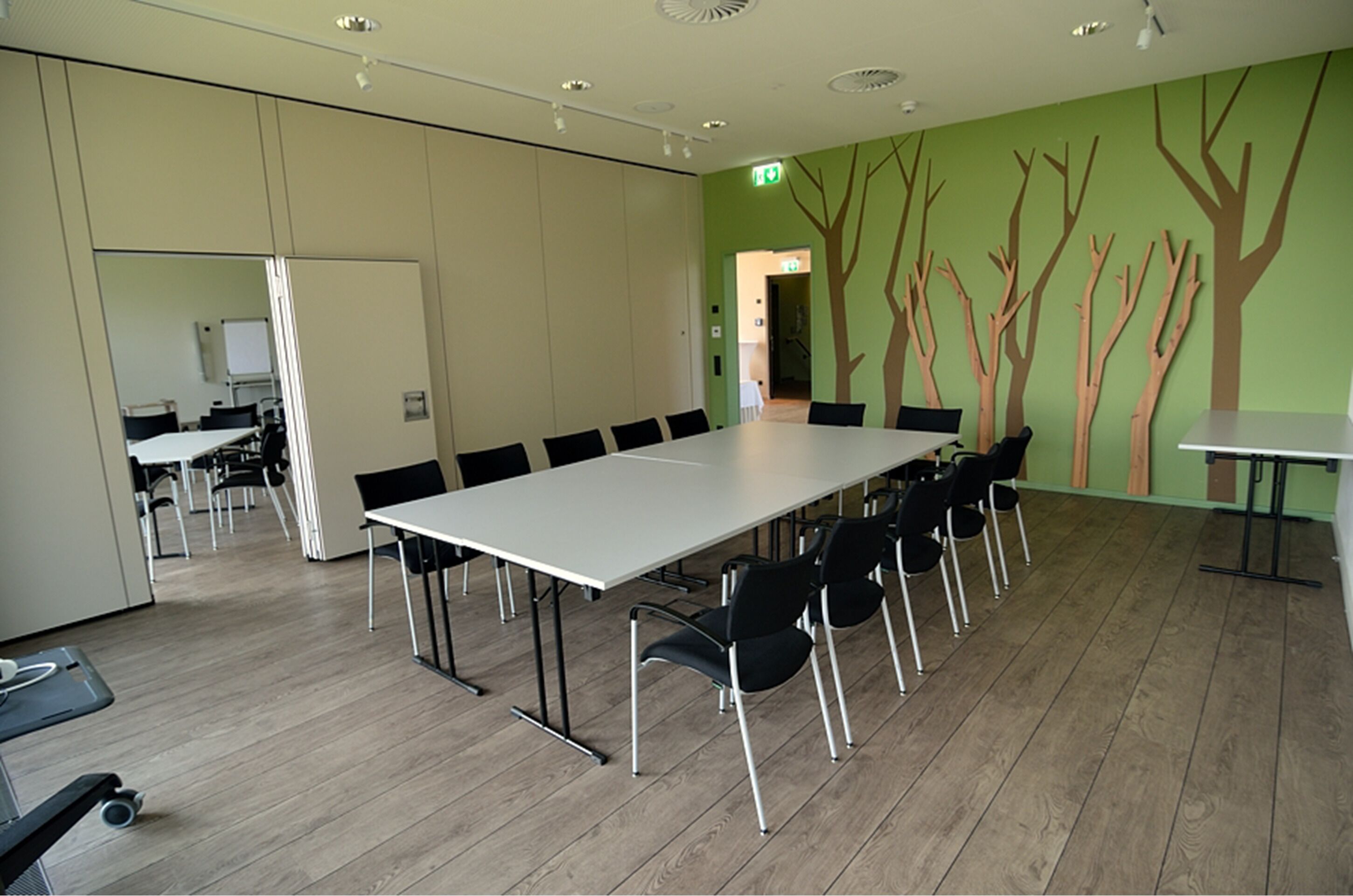 Ein Konferenzraum mit langem Tisch und Stühlen, einer Whiteboard und einer grünen Wand mit Baumdekorationen.