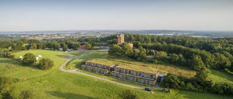 An aerial view of a modern building complex with multiple units, surrounded by lush greenery and a distant castle-like structure.
