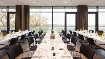 Un comedor con mesas dispuestas para una comida, ventanas amplias y vistas a un lago.