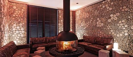 A modern lounge with a central wood-burning stove, leather sofas, and a unique wall design.